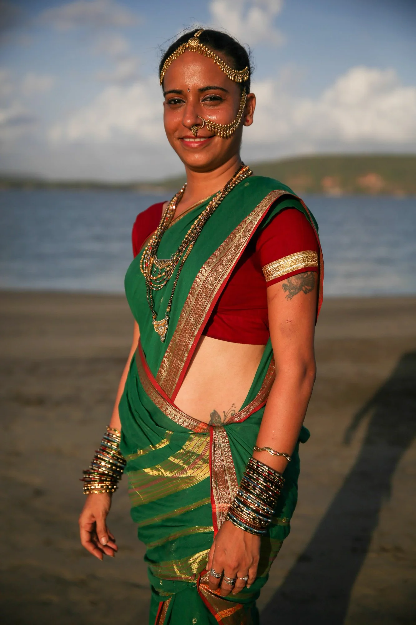 Smiling woman in a green and red sari with traditional gold jewelry standing by a lake