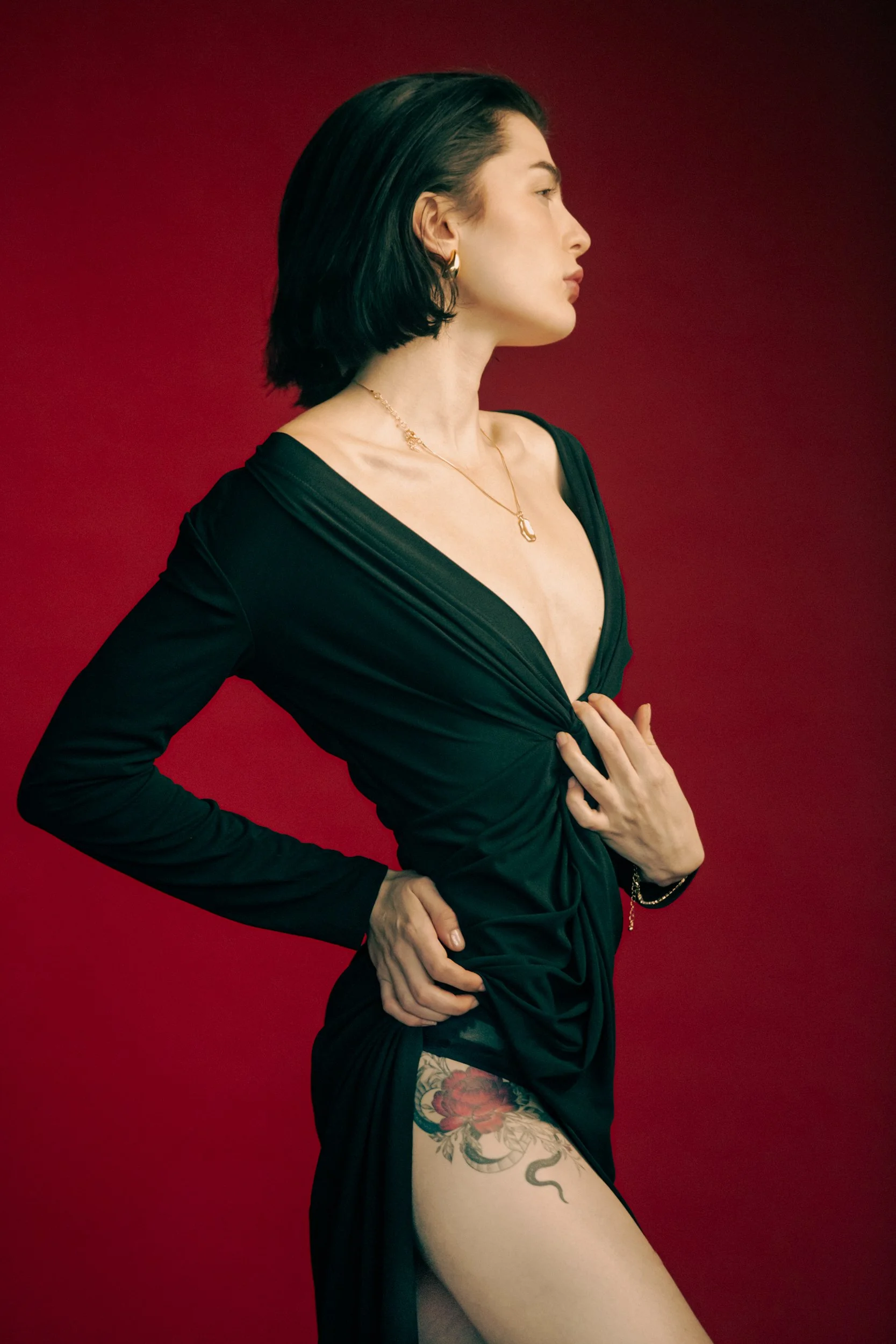 A woman in a black dress with red background, showing a tattoo on her thigh, gold jewelry, dark hair, and a side profile pose.
