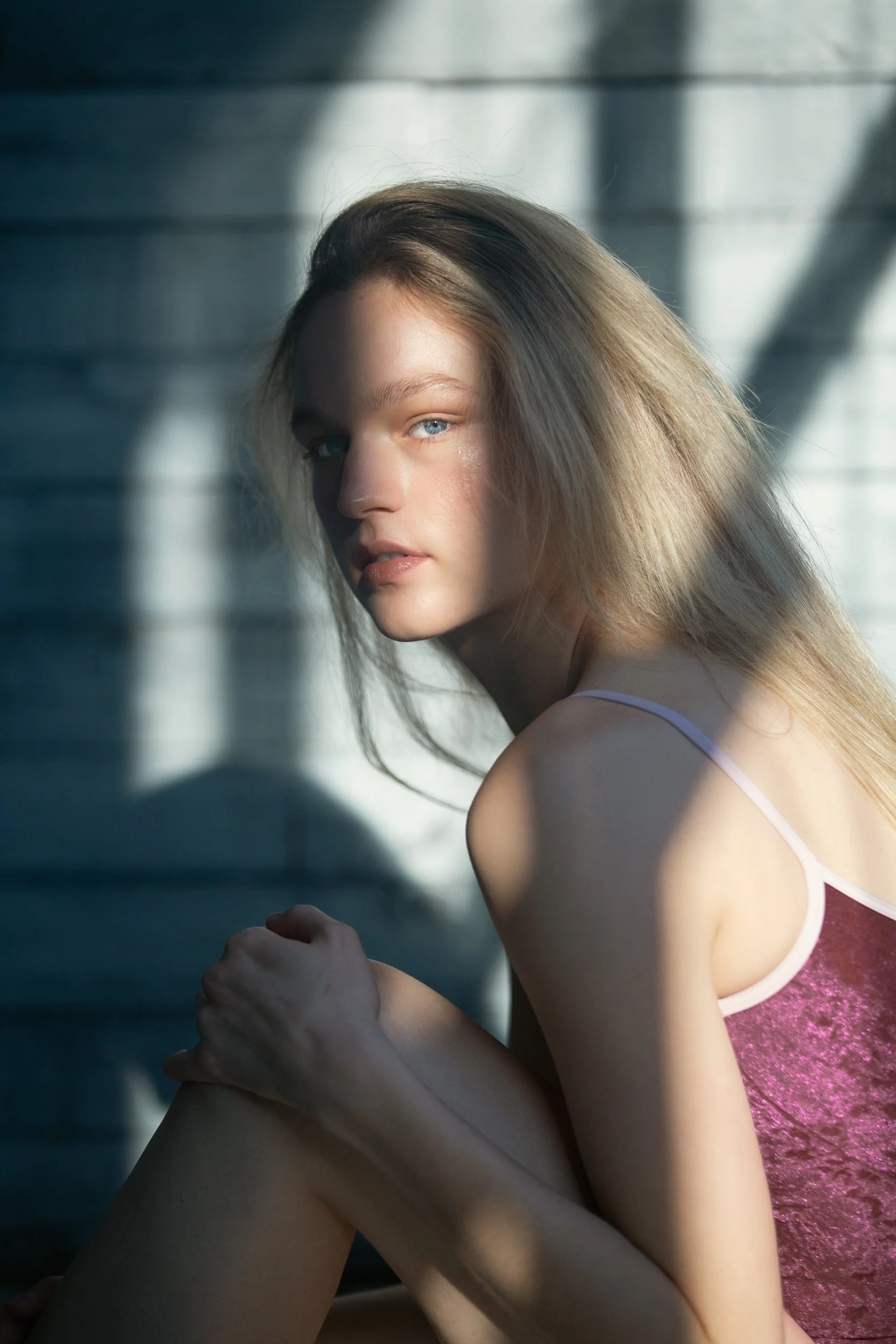 A young woman with long blonde hair and blue eyes looking at the camera, sitting in front of a wall with shadows cast on it, wearing a pink tank top.