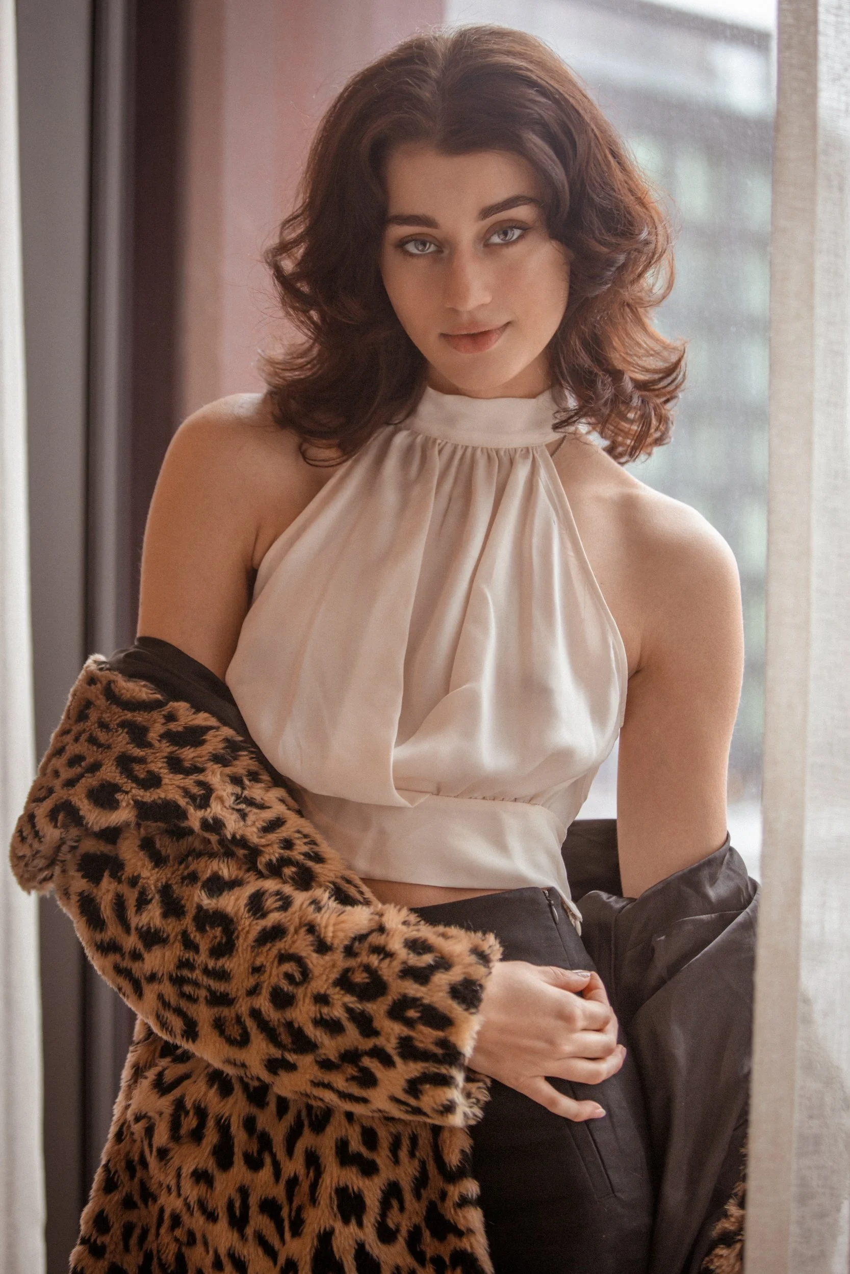 A woman with shoulder-length wavy brown hair and light skin, wearing a sleeveless cream-colored top and a leopard print coat, standing near a window.