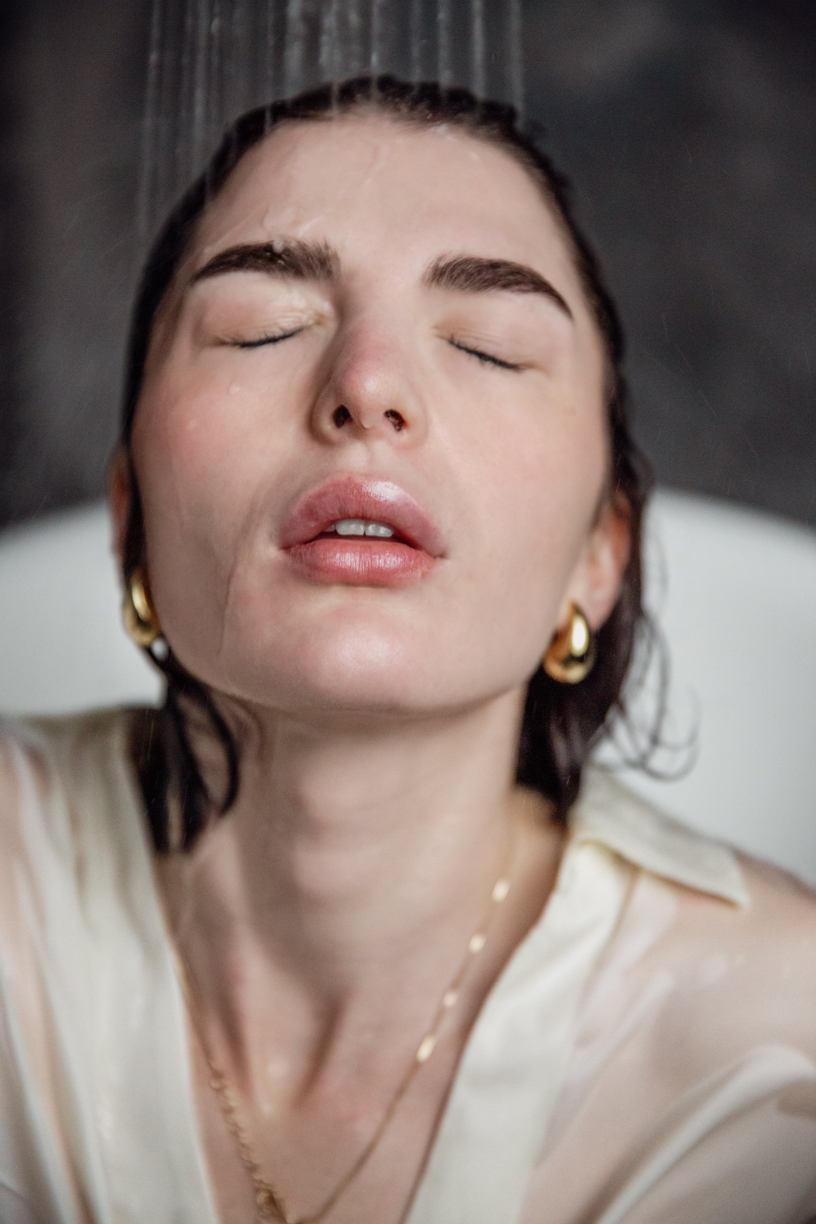Close-up of a woman with closed eyes, wet hair, and light skin, wearing gold earrings and a cream-colored blouse with a gold chain necklace.