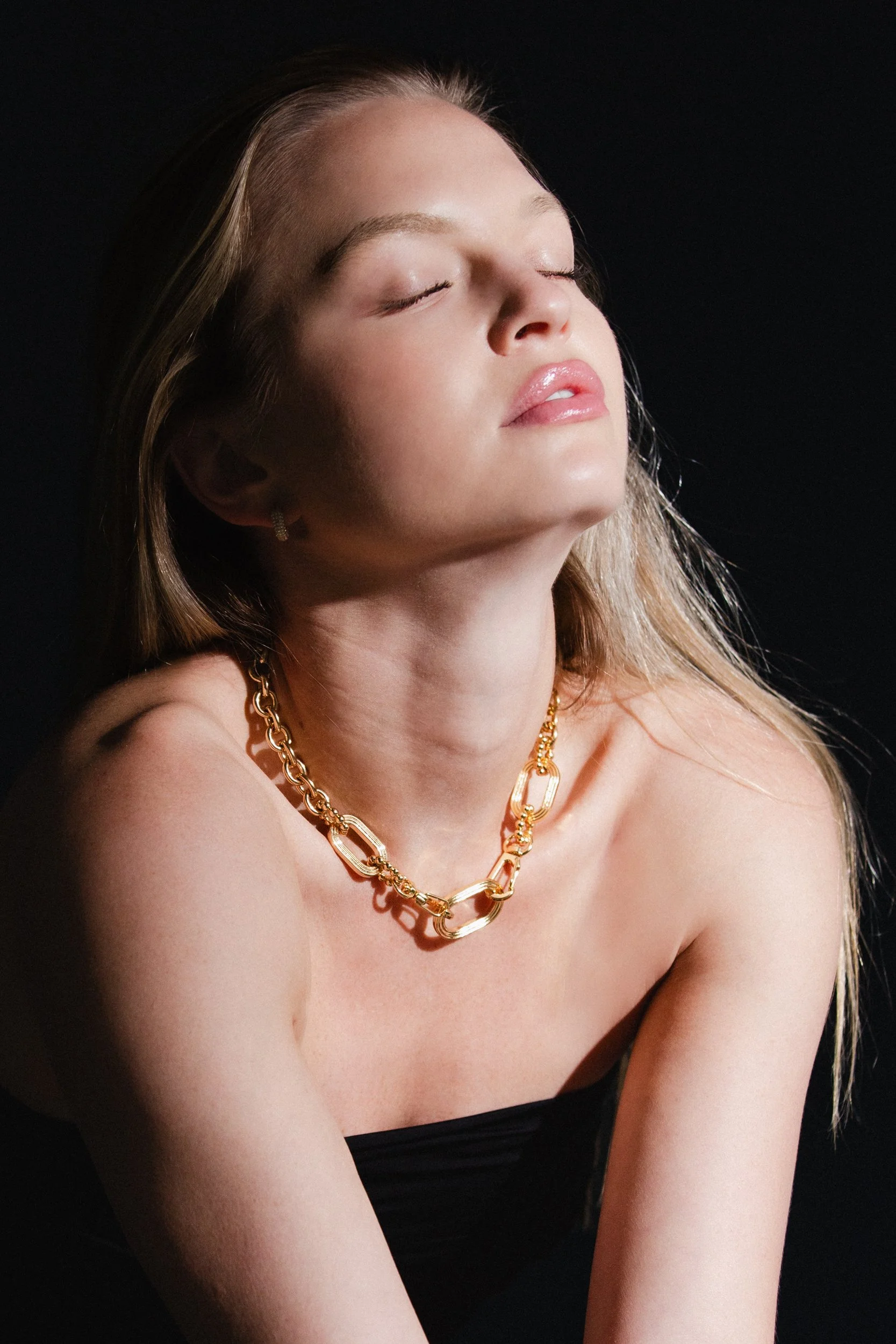 A woman with long blonde hair wearing a gold chain necklace and earrings, with her eyes closed and head tilted slightly back.