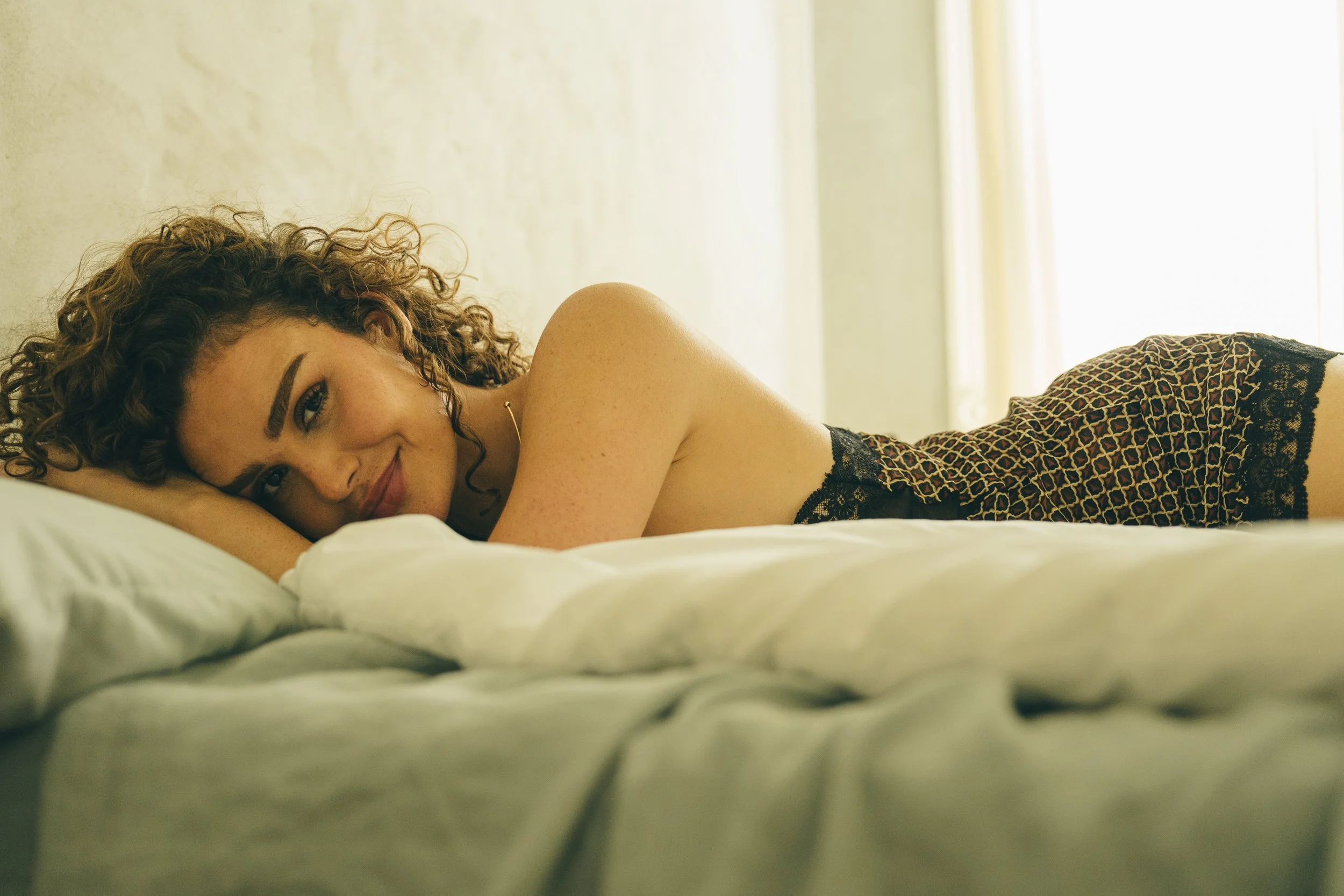 A woman with curly hair lying on a bed, smiling at the camera in a softly lit room.