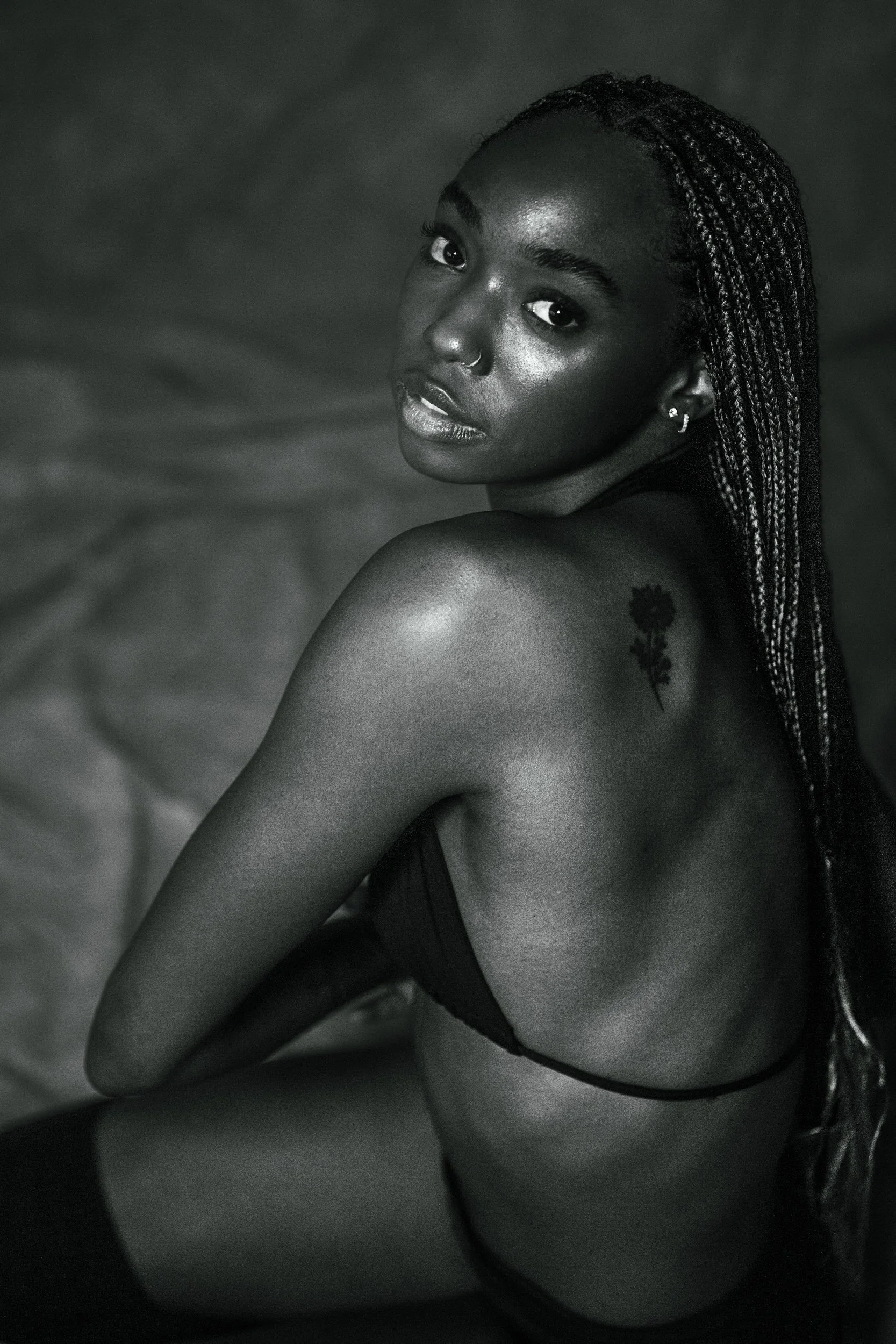 A black and white photograph of a young woman with braided hair, wearing earrings and a nose ring, looking over her shoulder with a serious expression. She has a tattoo of a sunflower on her shoulder and is dressed in dark clothing.