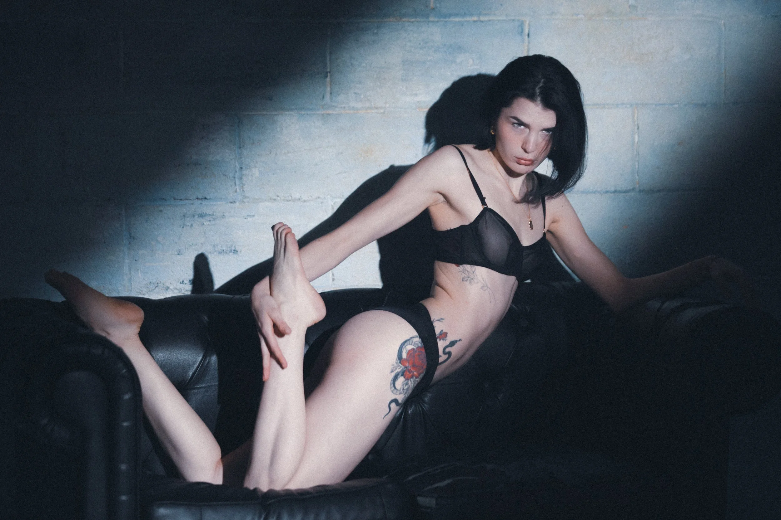 A woman with black hair and tattoos, dressed in black lingerie, is posing on a black leather couch against a gray brick wall, with dramatic lighting creating shadows.