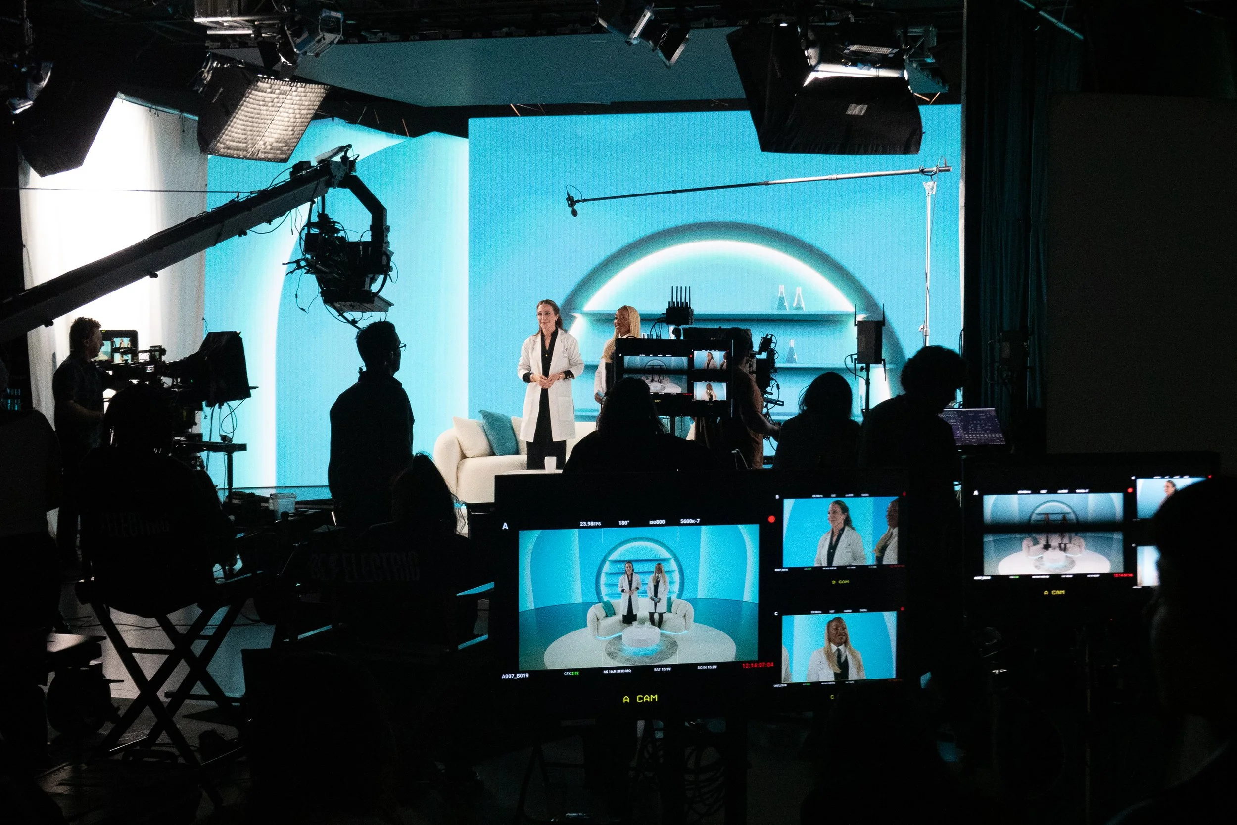 Behind-the-scenes view of a television or livestream production studio with multiple cameras, crew members, and large screens displaying a woman in a white coat on a set with a blue background.