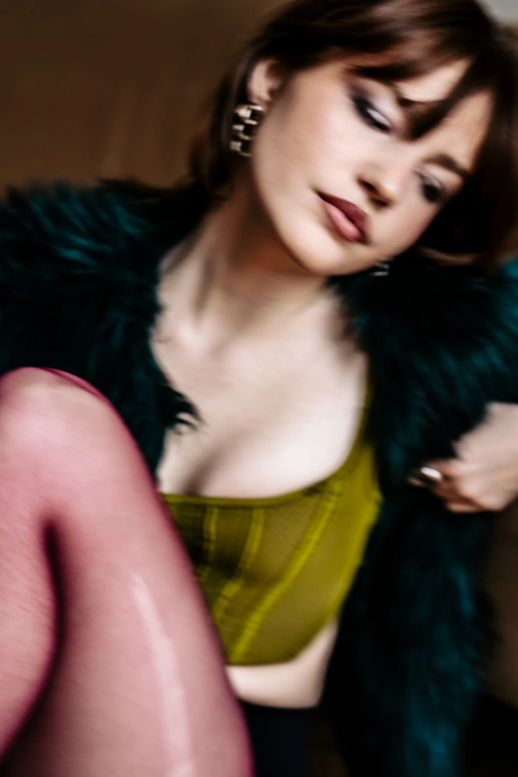 A woman with dark hair and earrings is looking downward, wearing a dark fur coat and a yellow-green top. In the foreground, a person's arm or leg is visible, wearing pink or peach-colored clothing. The background is blurred.