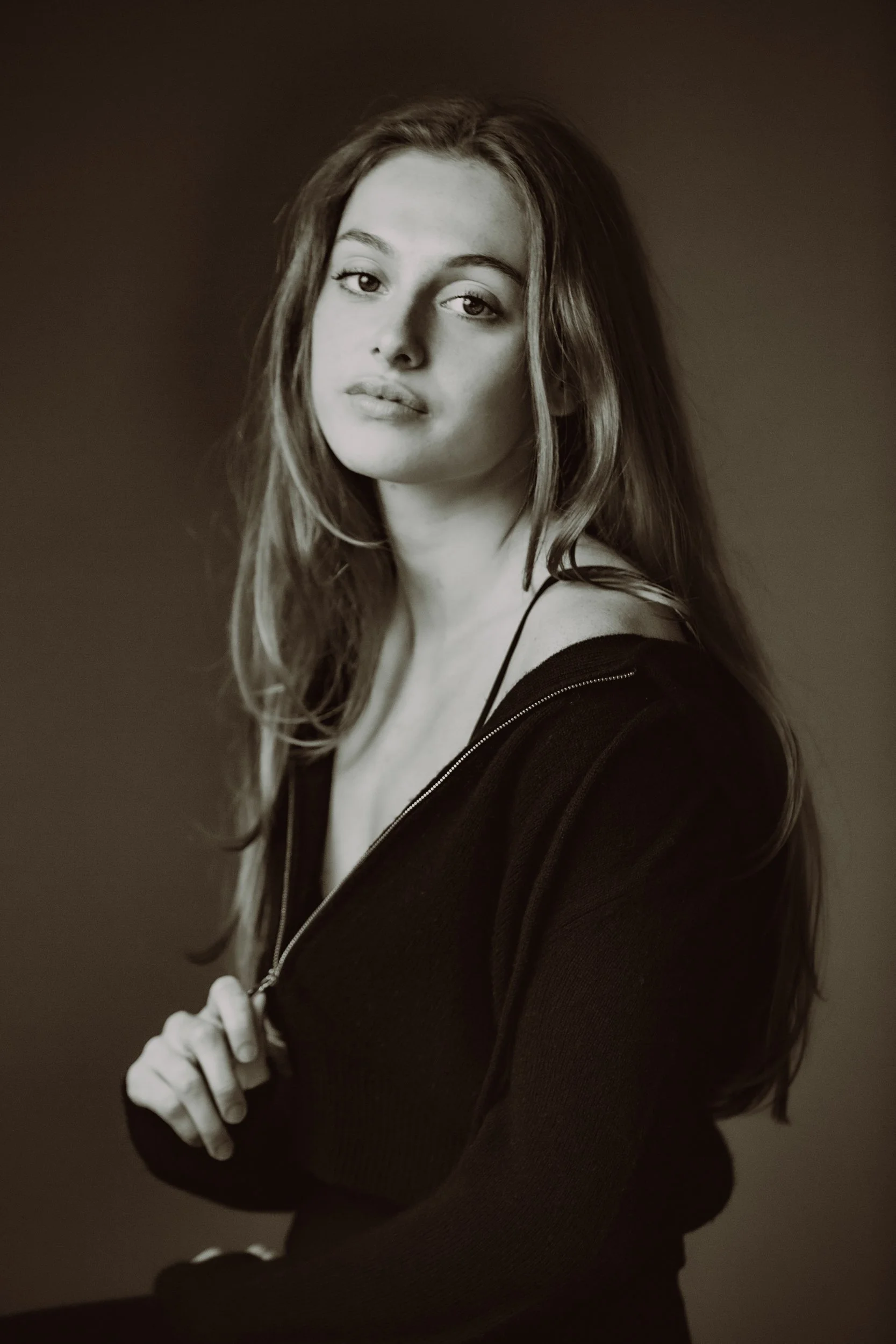 Black and white portrait of a young woman with long hair, wearing a dark top with thin straps, standing against a plain background.