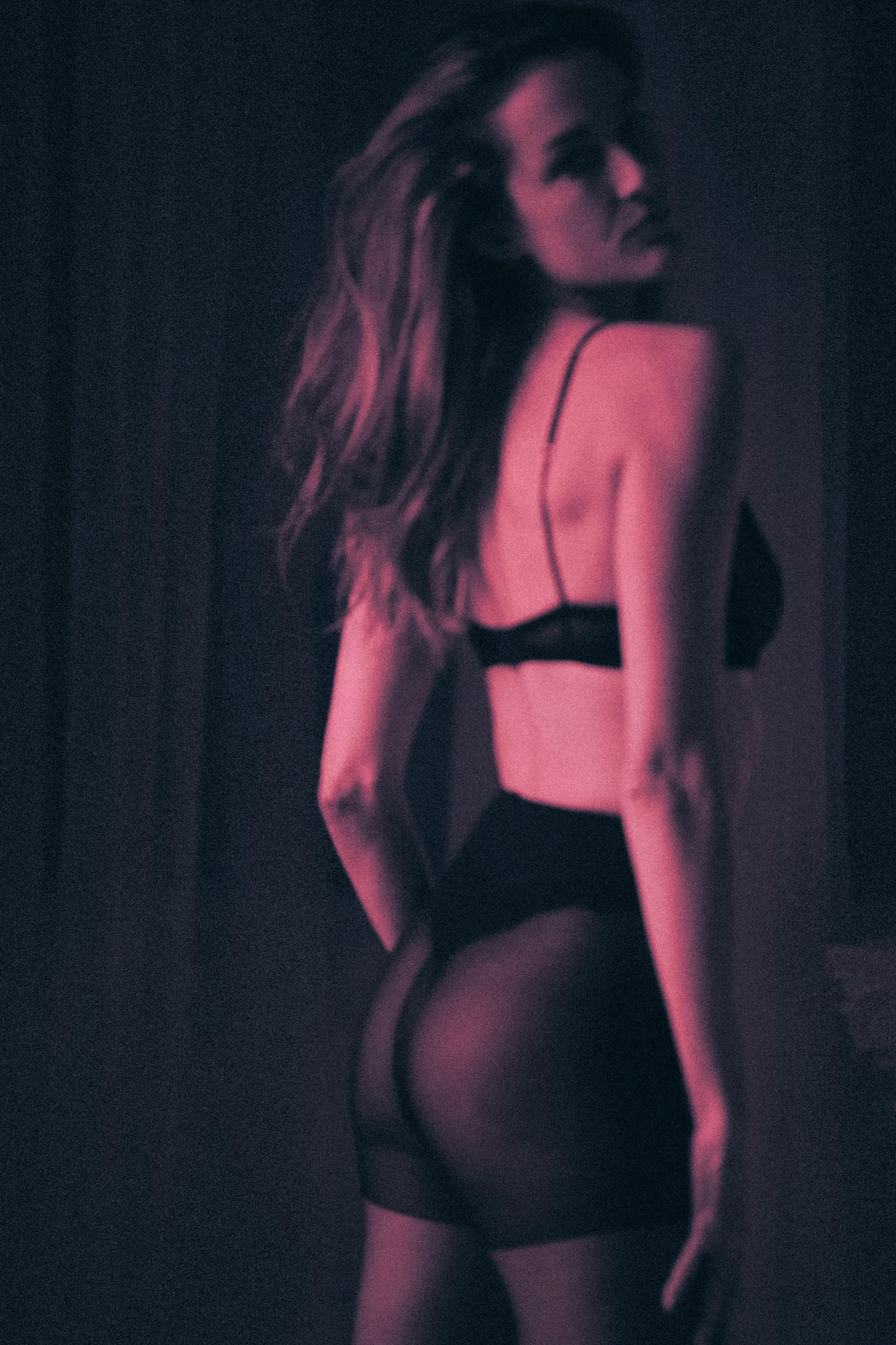 A woman with long hair wearing black lingerie and thigh-high stockings, photographed in a dark, moody setting with pinkish-purple lighting.