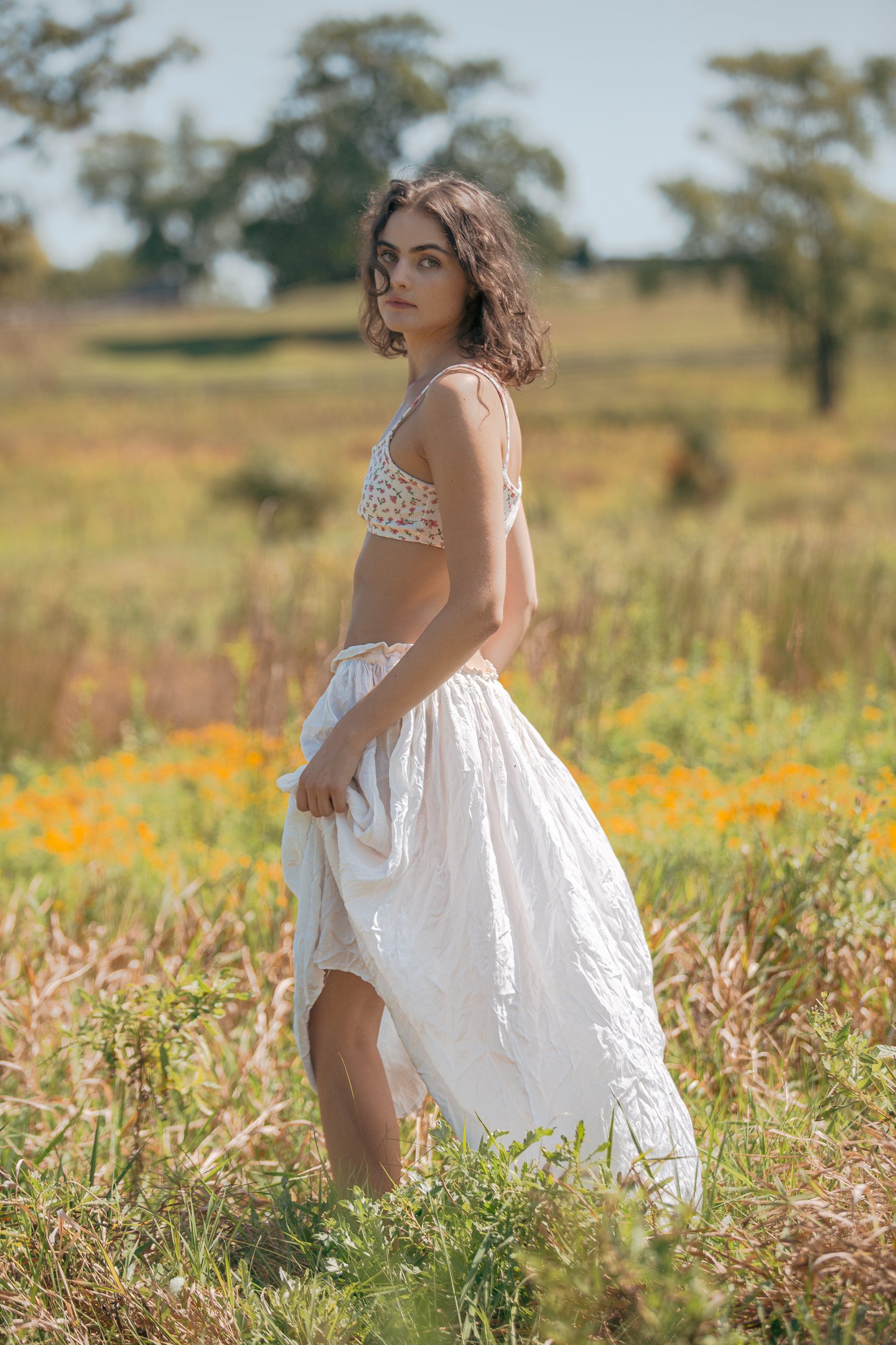 A young woman standing in a field of tall grass and orange flowers, wearing a floral crop top and a white flowing skirt.