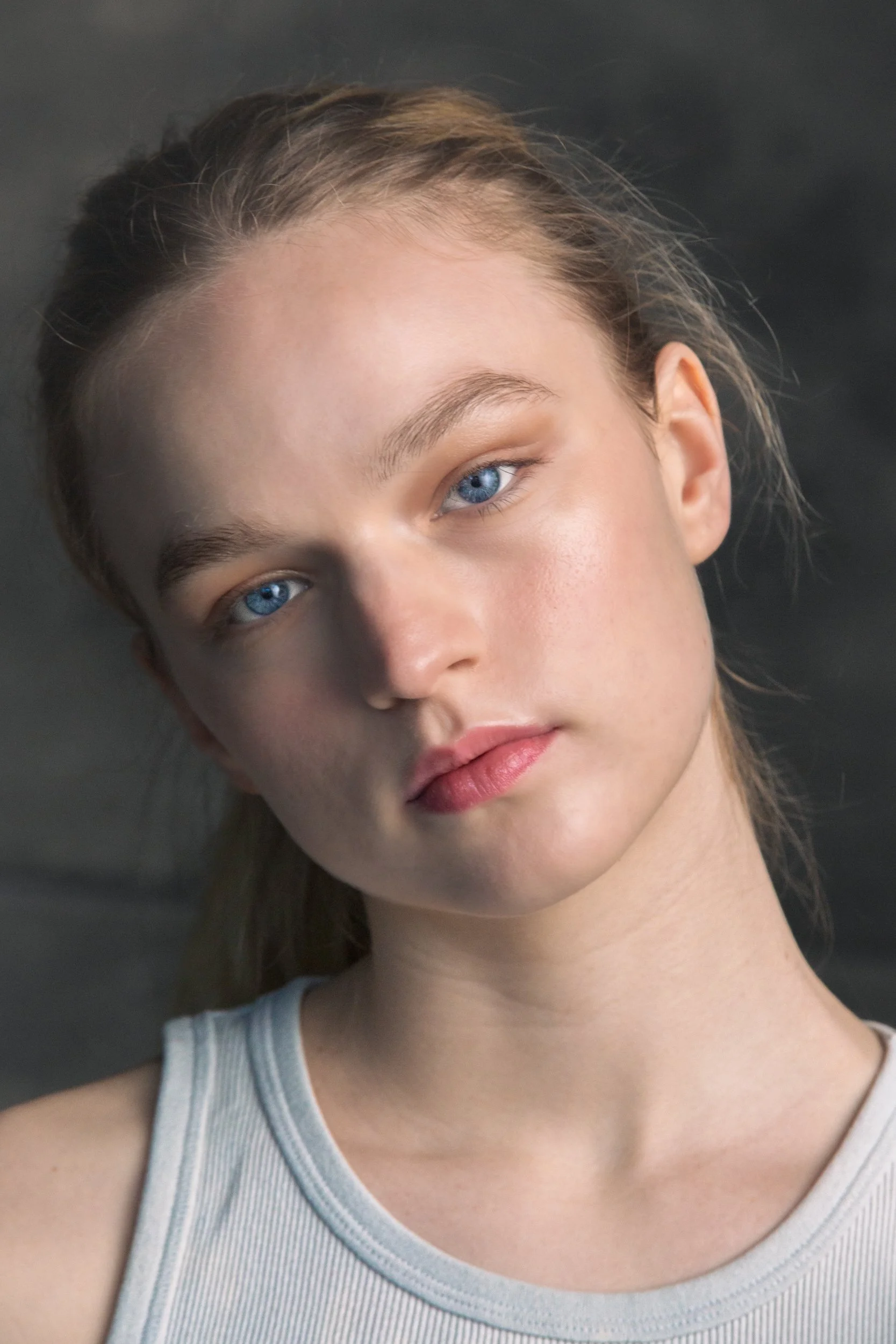 Close-up of a young woman's face with blue eyes, light skin, and brown hair, wearing a gray tank top, against a dark background.