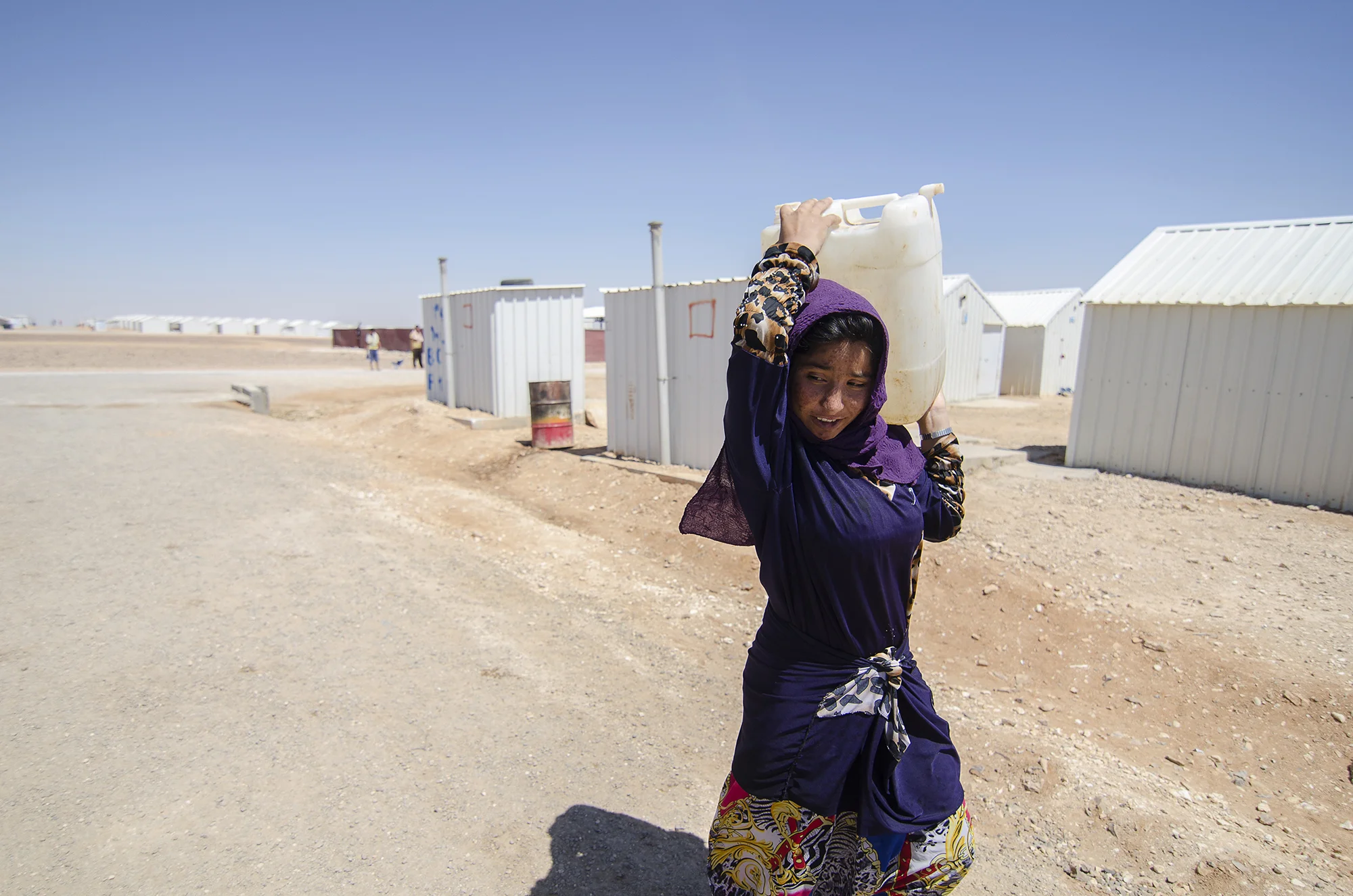 A refugee carries water back to her family's caravan in Jordan's Azraq Refugee Camp for Syrians, opened May 2014 in the country's eastern desert province and camp namesake, Azraq. Unlike Zaatari's chaotic, impromptu inception, Azraq's development was preplanned by the UNHCR as an humanitarian response to increasing numbers of refugees. See more at Al Jazeera. June 2015.