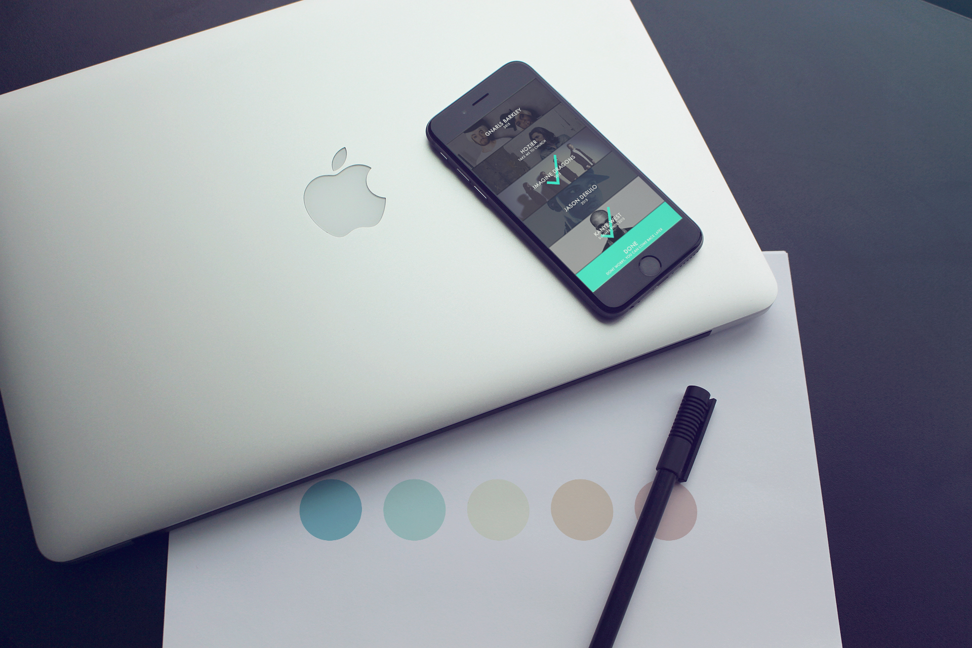 A closed silver MacBook laptop, a black iPhone with an app open on the screen, a black pen, and a color palette sheet with five different colored circles on a dark surface.