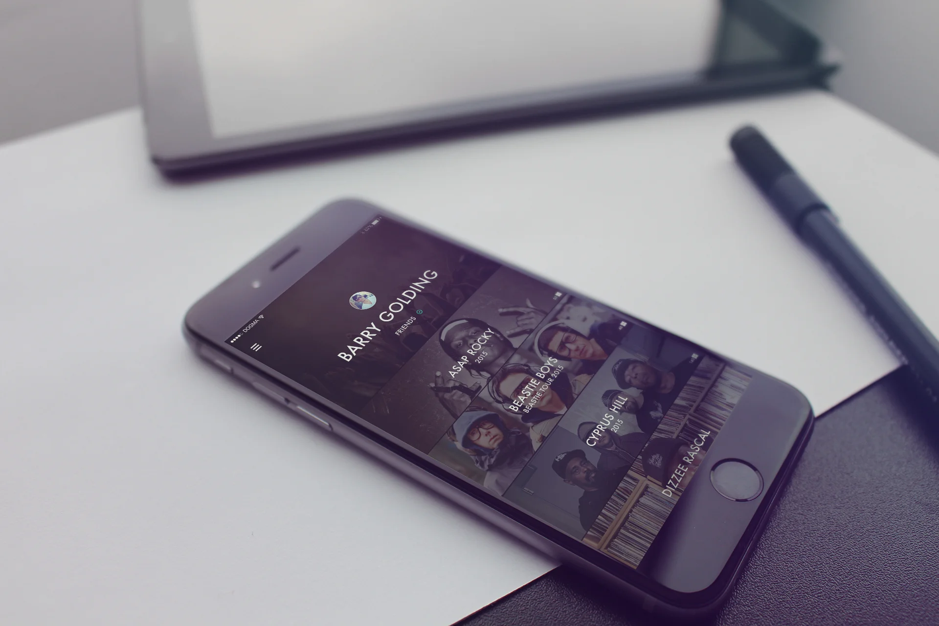 An iPhone showing a music app with Barry Golding as the artist, placed on a white table with a black pen nearby and a silver laptop in the background.