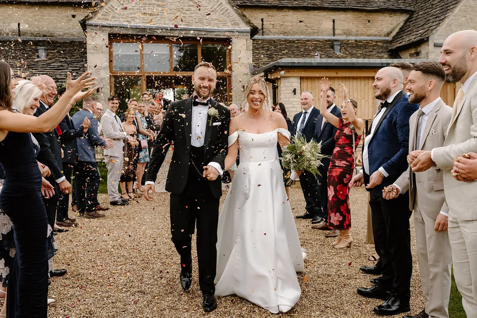 A newly married couple walks hand in hand through a confetti celebration, surrounded by friends and family outside a rustic building.