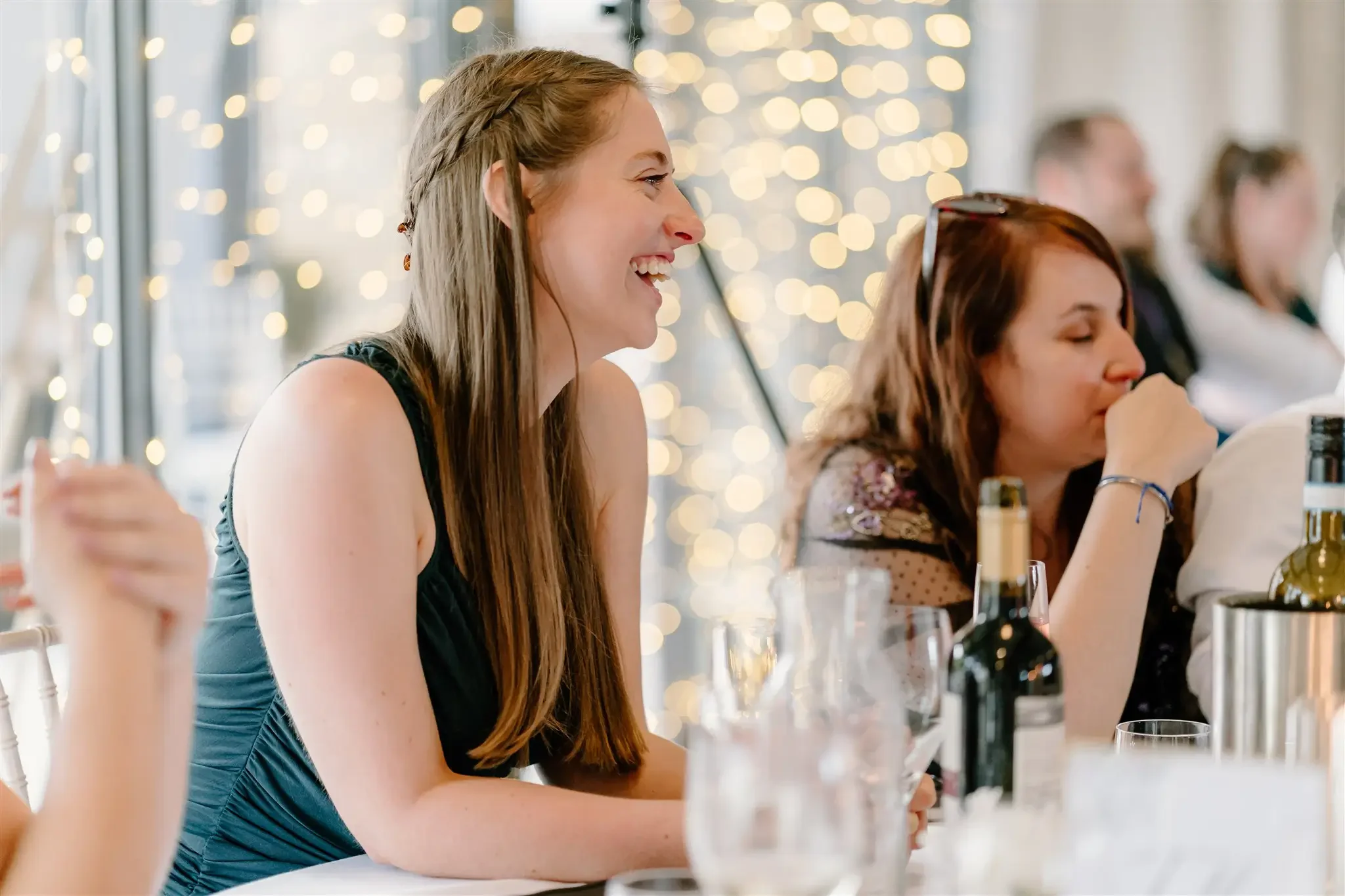 Two women sitting at a table laughing and talking at a social gathering, with blurred lights in the background.