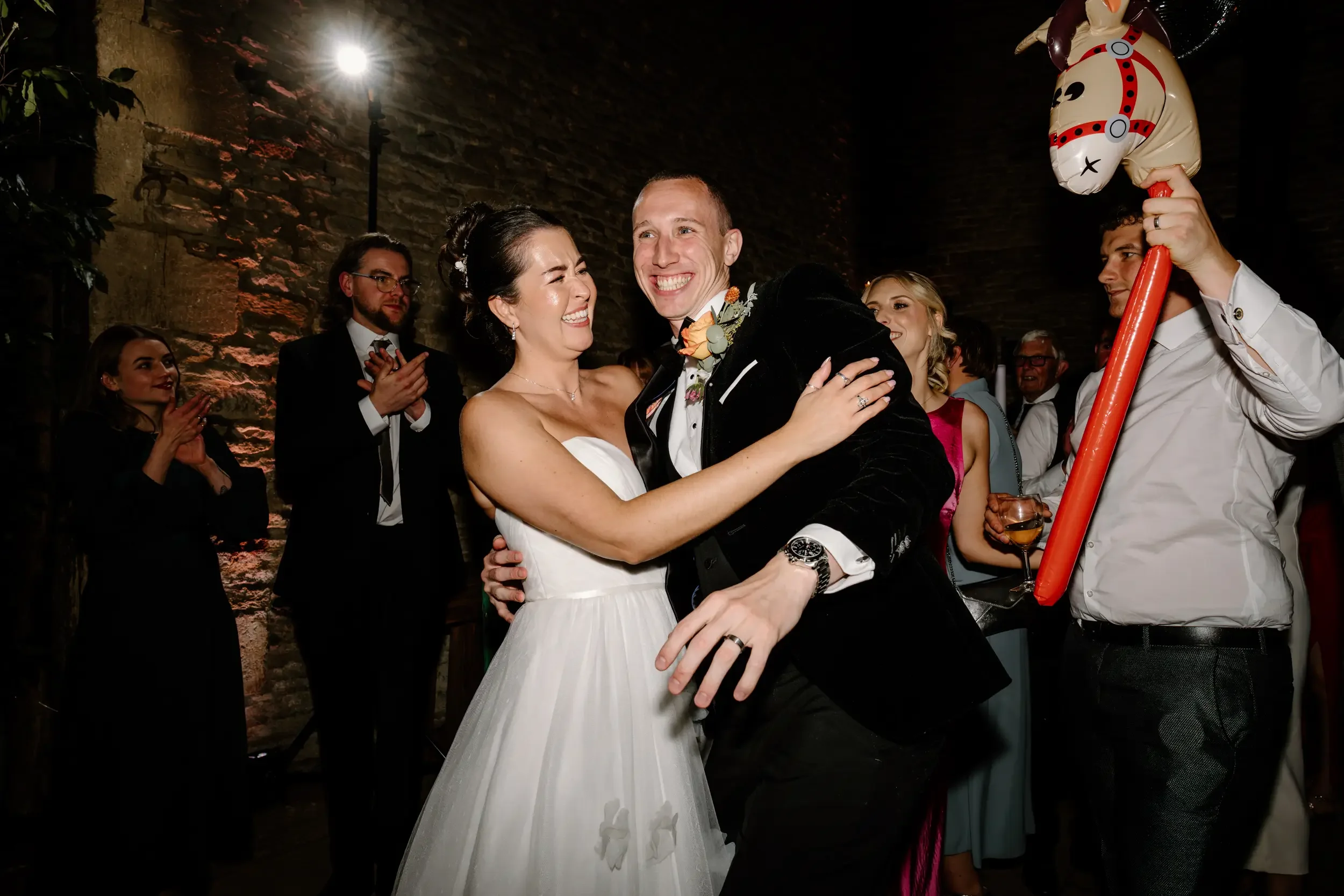 A bride and groom are dancing and smiling at their wedding reception, surrounded by guests who are clapping and watching.