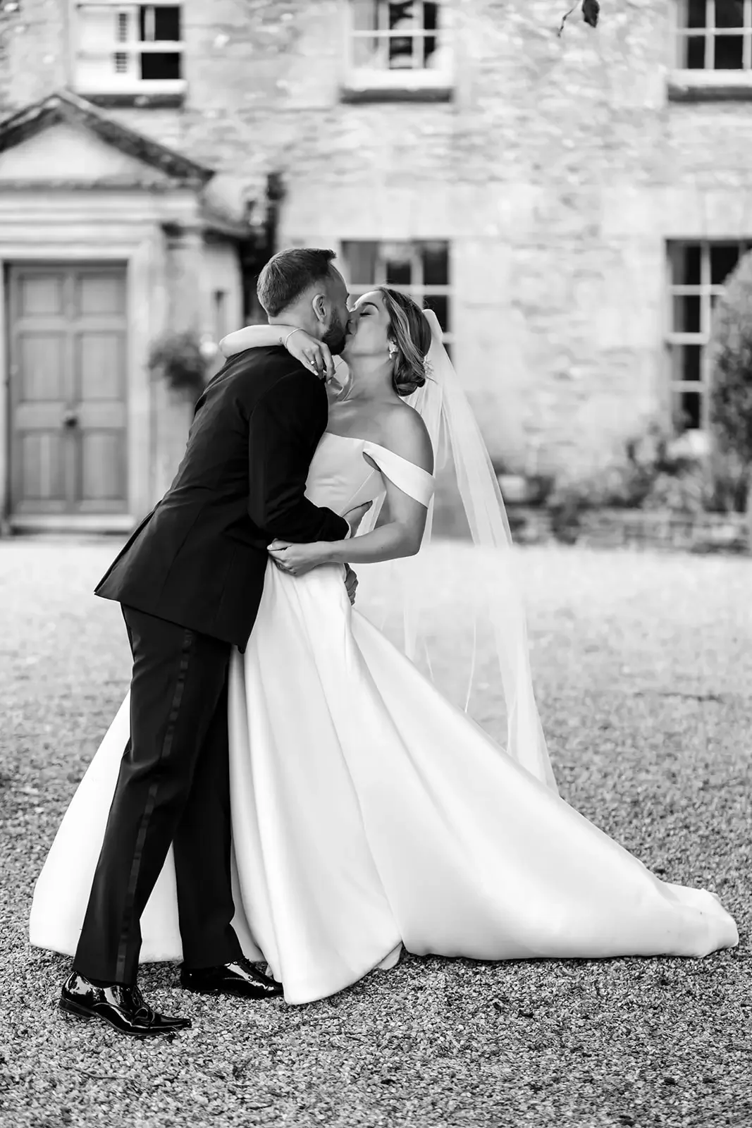 A black-and-white photo of a bride and groom sharing a kiss outdoors, with a rustic brick building in the background. The groom is in a dark suit, and the bride is in an off-shoulder wedding gown, with a veil.
