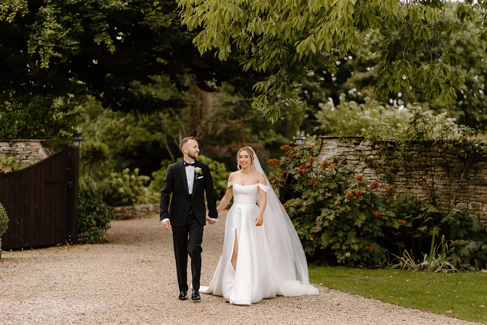 A bride and groom are walking hand in hand outdoors on a gravel path, surrounded by green trees and bushes, with a stone wall in the background.