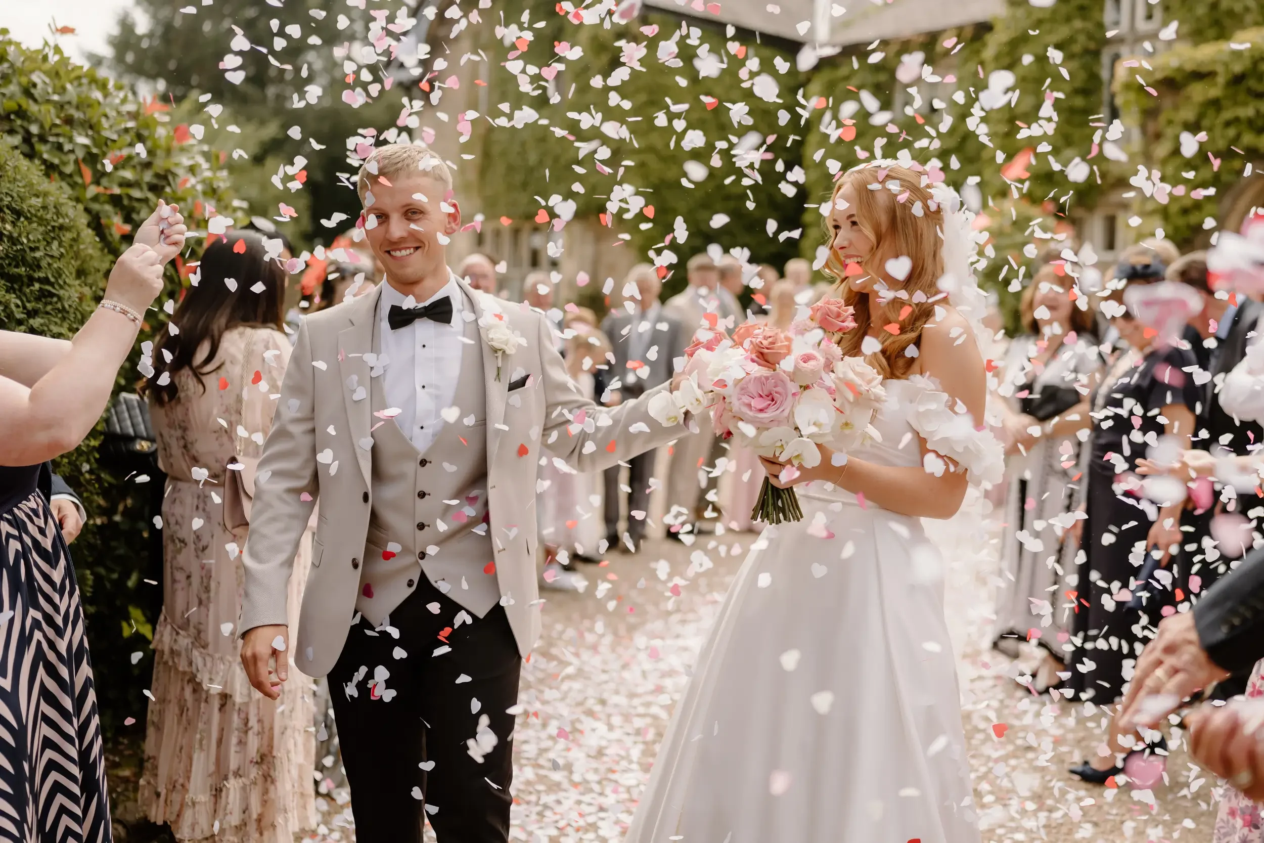 A newlywed couple walking hand in hand through a shower of heart-shaped confetti, surrounded by cheering guests at an outdoor wedding ceremony.