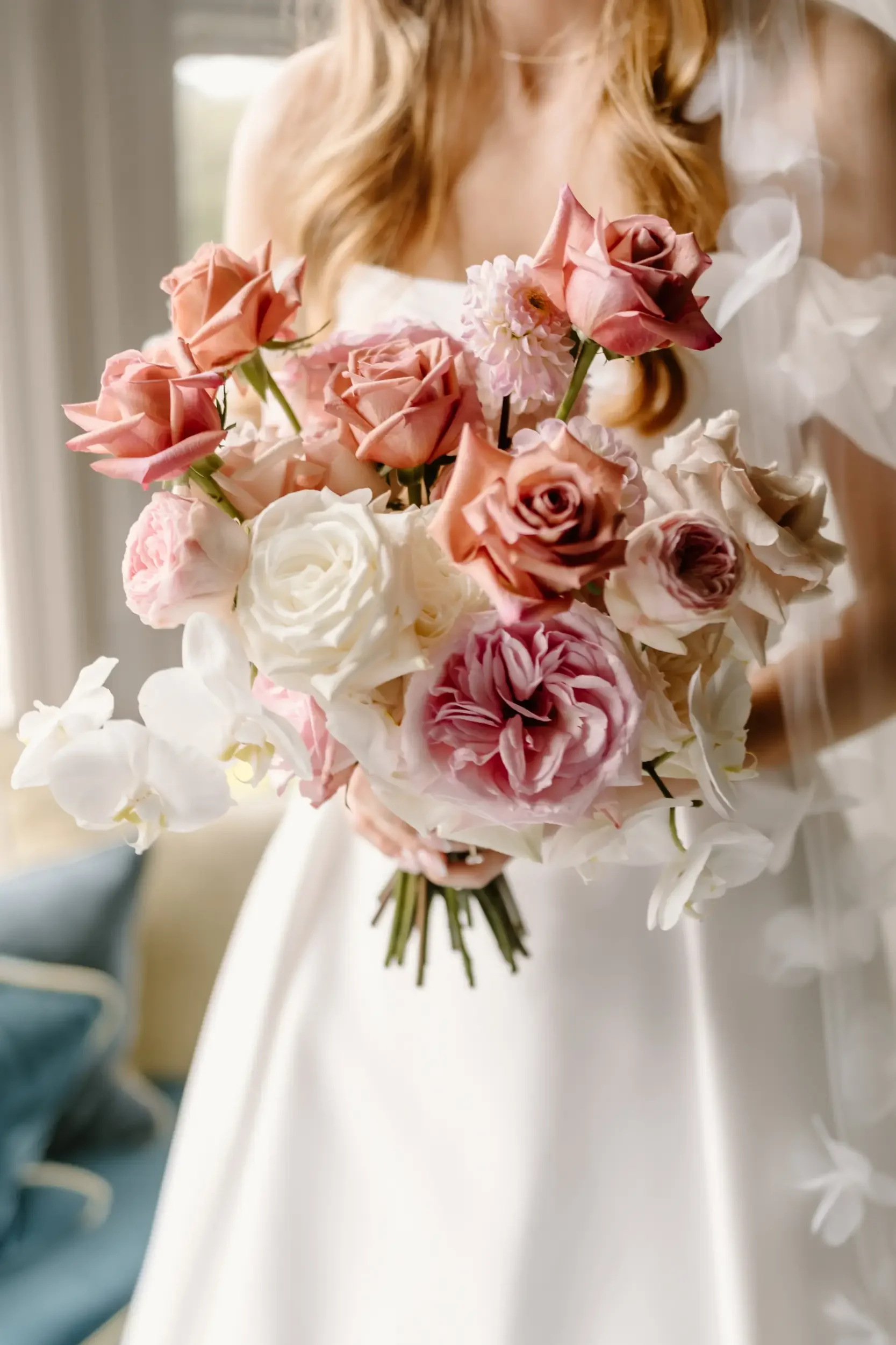 A woman in a white dress holding a bouquet of pink, white, and peach roses, with some white orchids, in front of a window.