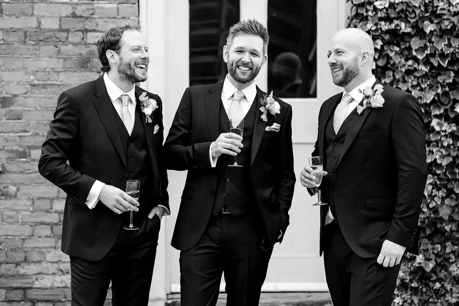 Four men dressed in tuxedos standing outside, holding glasses of champagne, smiling and conversing during a celebration or wedding.