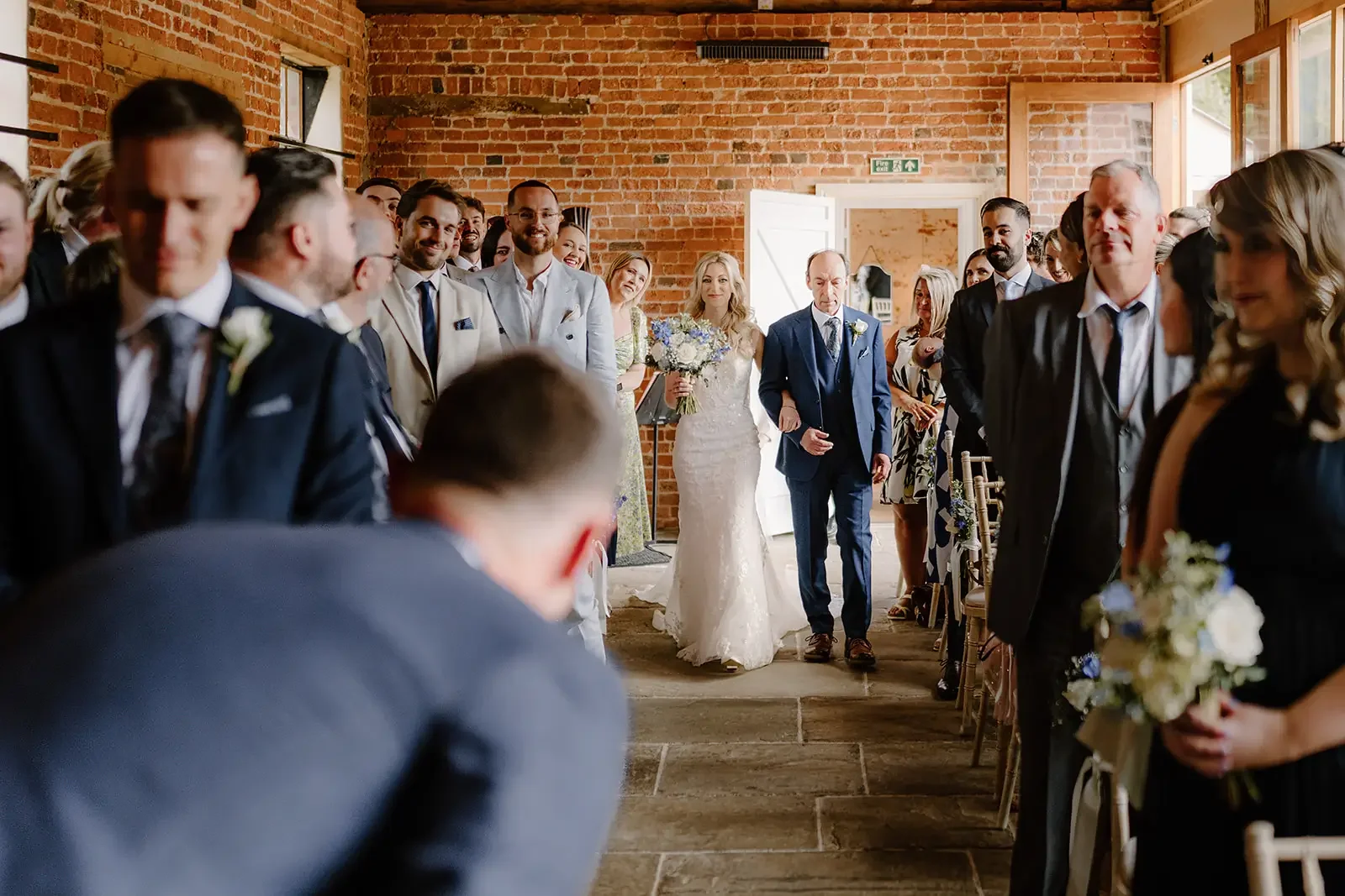Bride walking down the aisle with her father at a wedding ceremony inside a brick-walled venue, surrounded by seated guests.