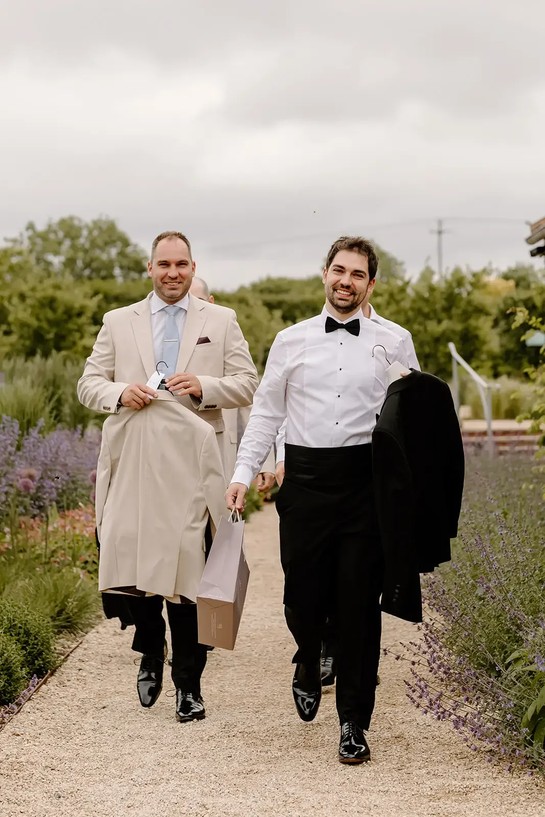 Two men in formal attire walking in a garden. One man is wearing a white tuxedo with a black bow tie and holding a black suit jacket. The other man is in a beige suit and beige coat, holding a hanger with a beige coat. Both are smiling.