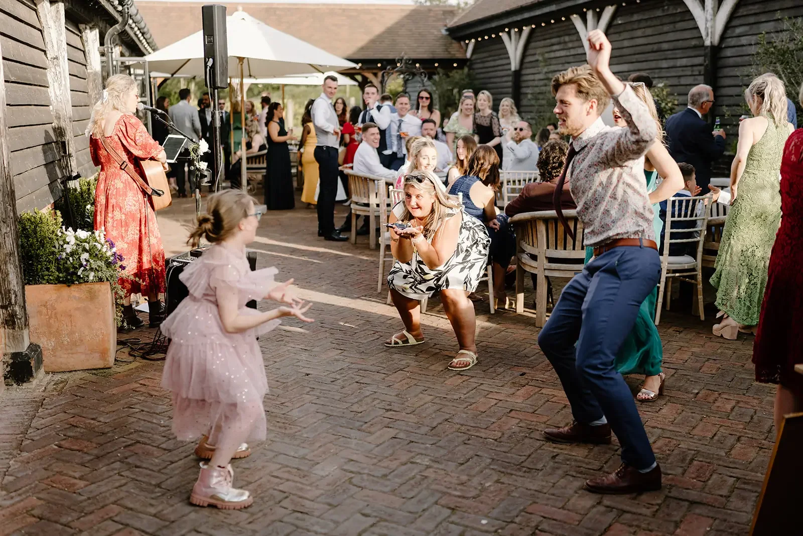 People dancing and enjoying music at an outdoor celebration or wedding reception, with a singer playing guitar on the left and guests seated at tables in the background.