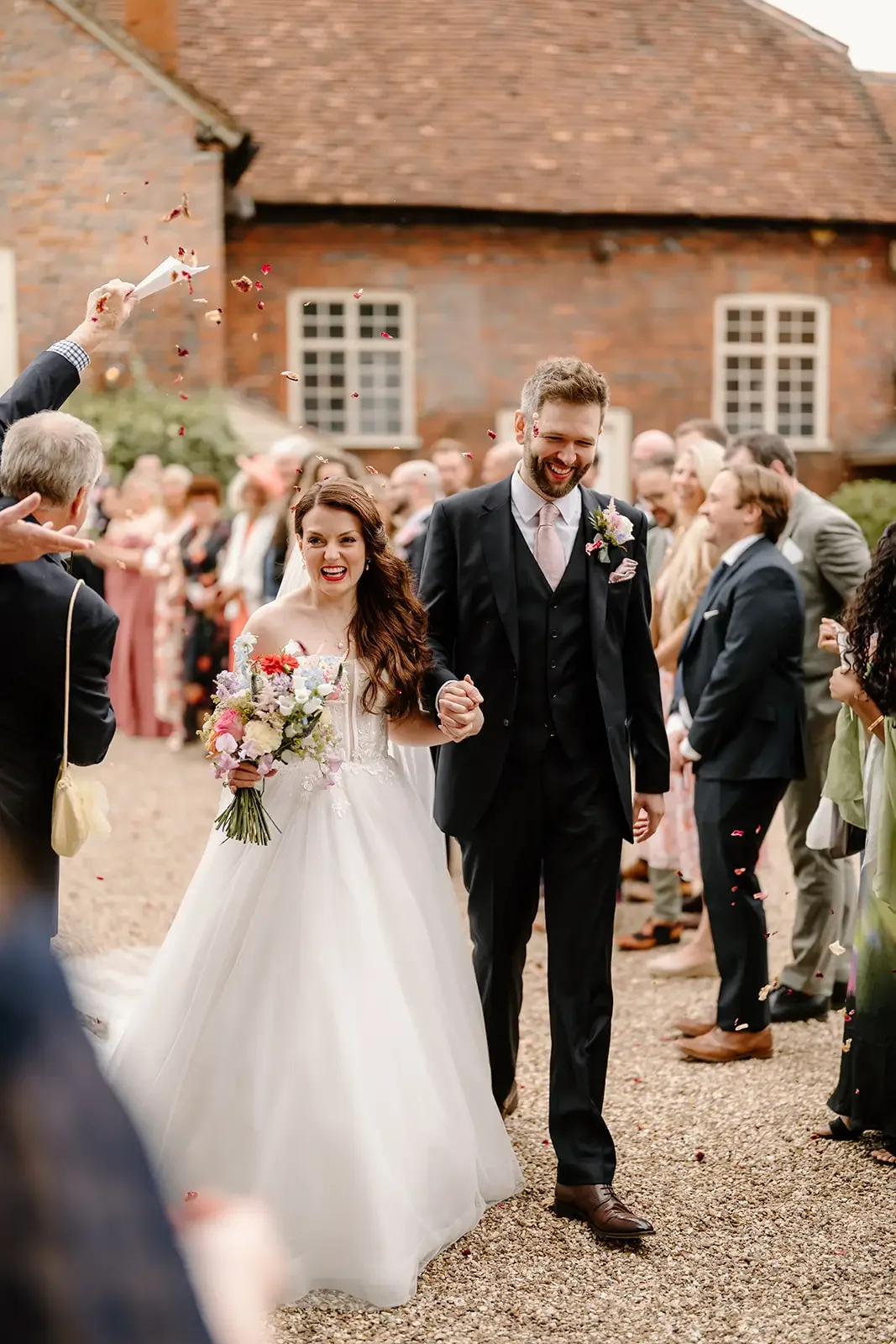 A newly married couple walking outside, holding hands, being showered with flower petals by wedding guests. The bride is in a white wedding dress holding a bouquet, and the groom is in a black suit with a pink tie.