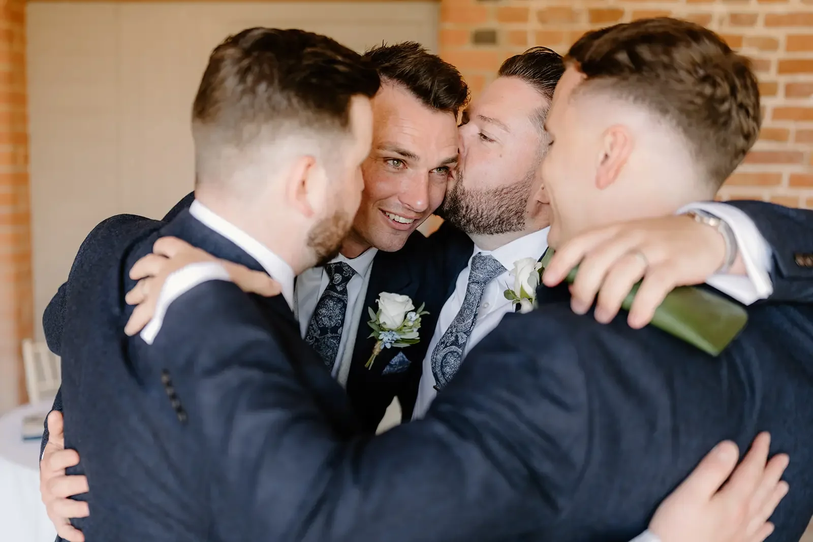 Four men in suits hugging and sharing a kiss on the cheek at a wedding, indoors with brick wall background.