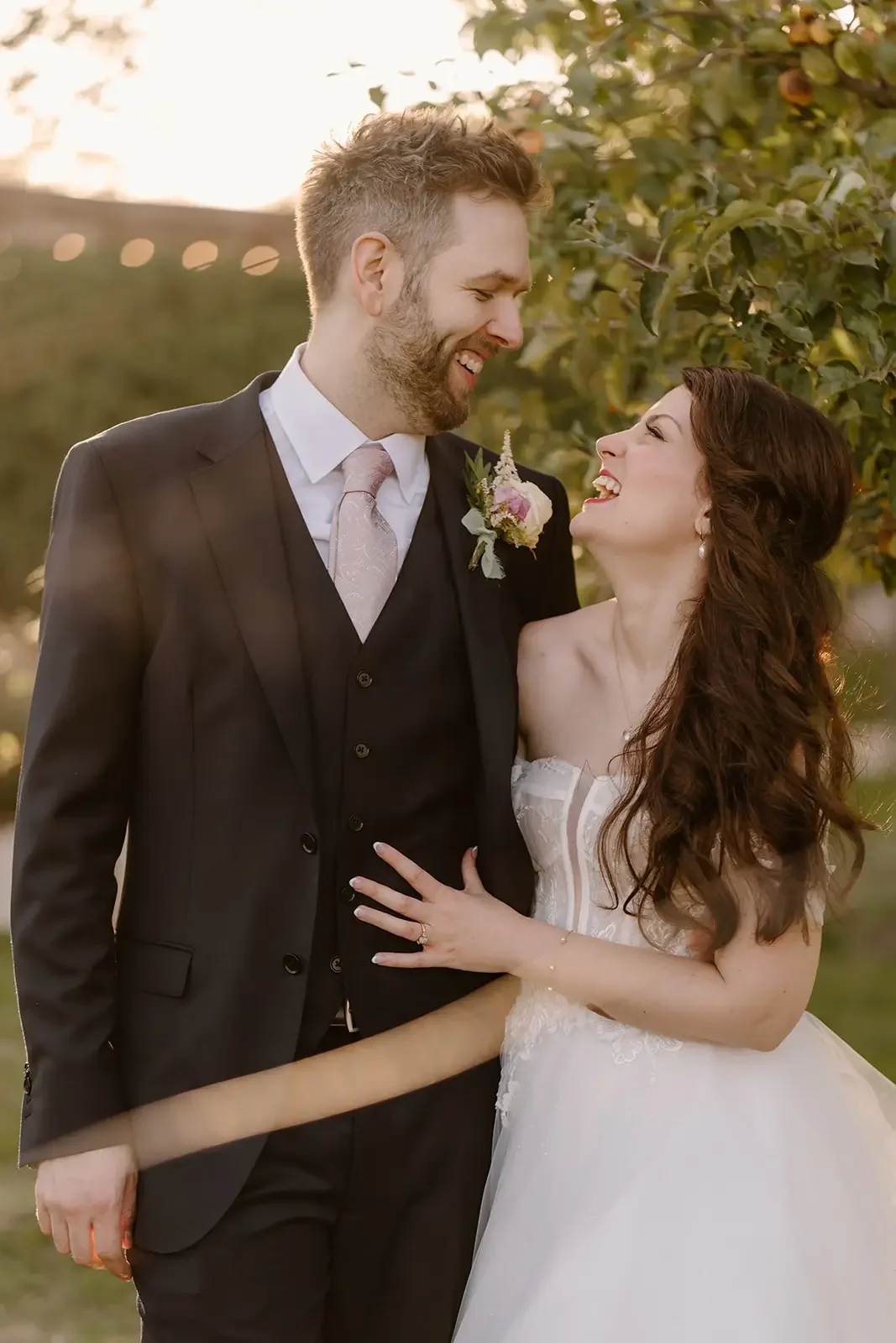 A happy couple in wedding attire sharing a joyful moment outdoors, with the groom in a suit and the bride in a white wedding dress, surrounded by greenery.