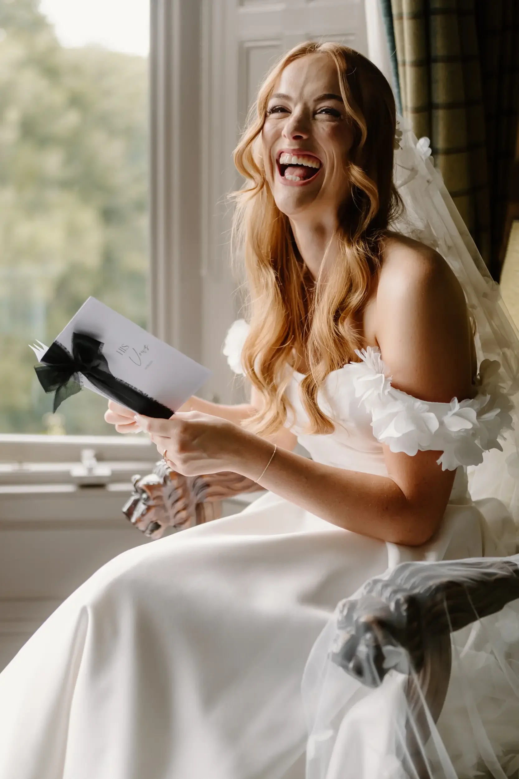 A smiling woman in a wedding dress sitting by a window, holding a card with the words 'Mrs. Love' written on it, with a joyful expression on her face.