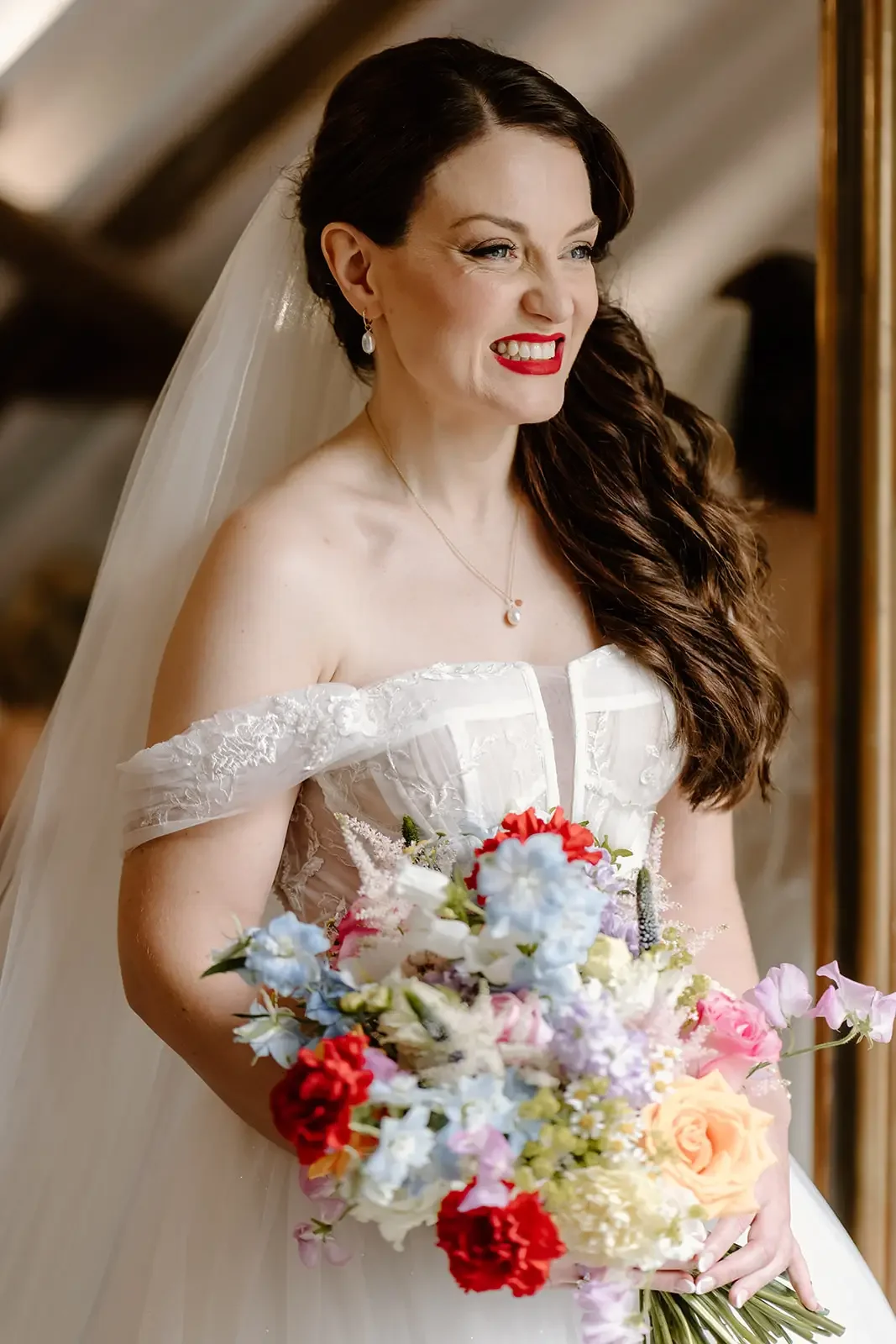 A bride with dark brown wavy hair, wearing red lipstick, pearl earrings, a pendant necklace, and a white off-the-shoulder wedding dress holding a colorful bouquet of flowers.