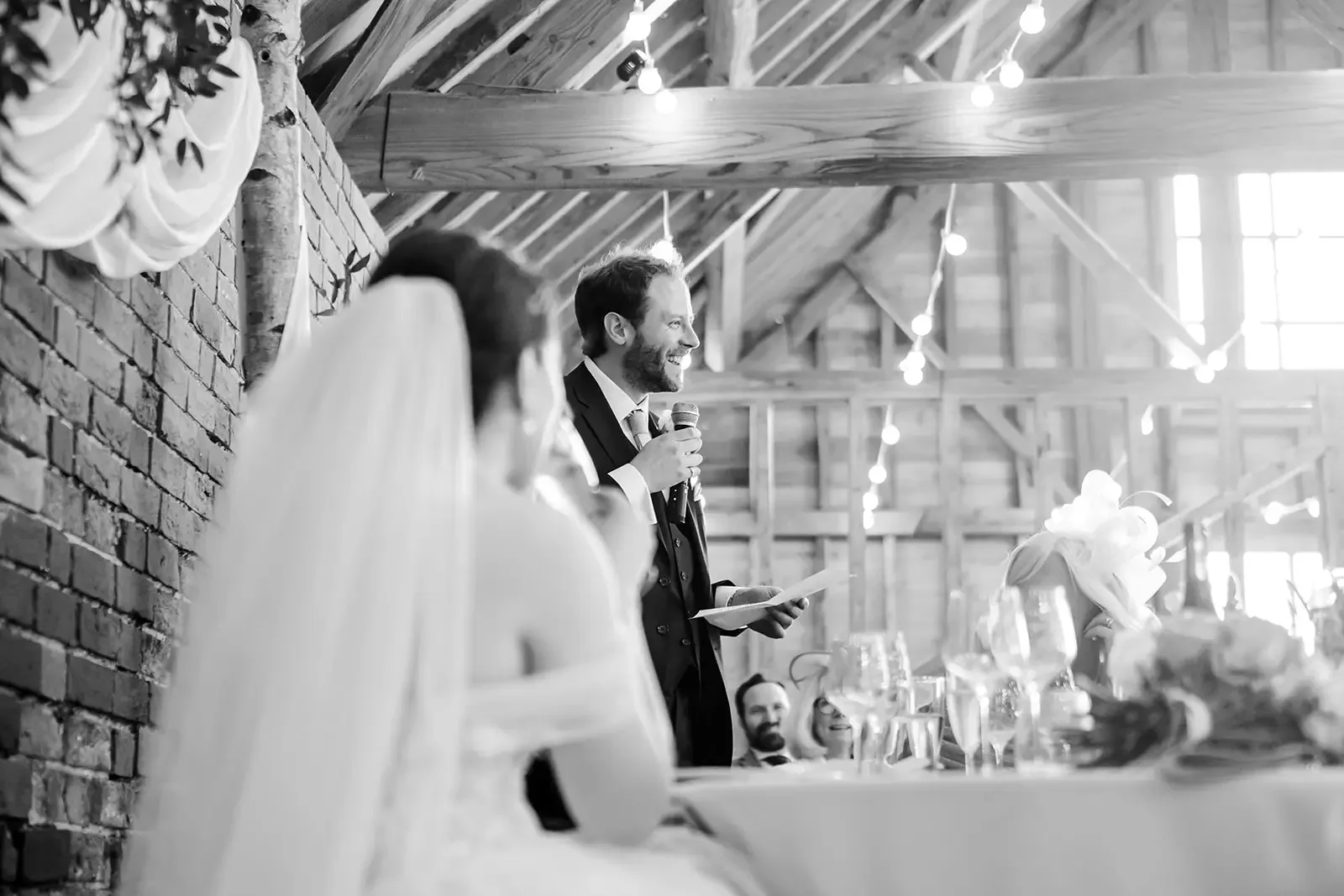 A man giving a speech at a wedding reception, with a bride in the foreground and other guests seated at a decorated table, in a rustic venue with string lights hanging from wooden beams.