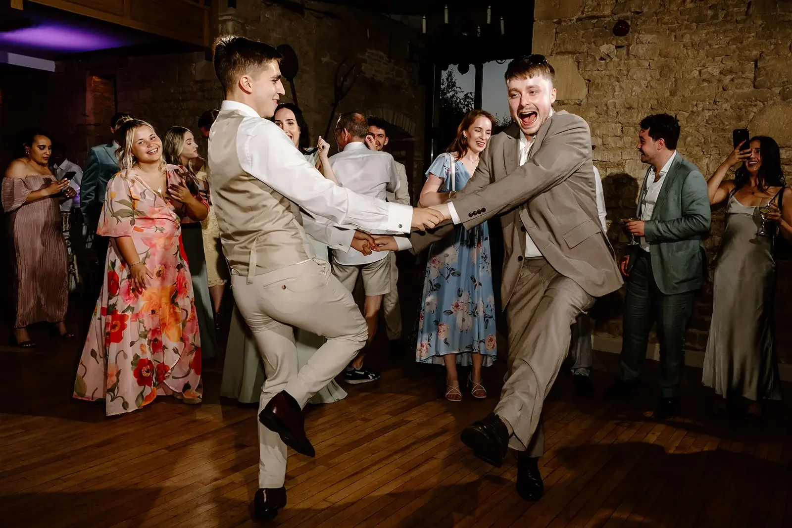 People dancing and celebrating at a wedding reception, with two men in suits holding hands and whirling on a wooden dance floor, surrounded by guests in formal and floral dresses.