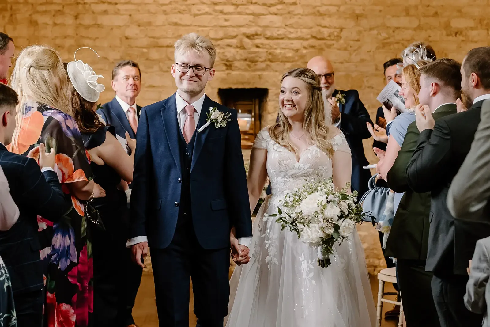 Bride and groom holding hands, surrounded by wedding guests in a rustic indoor setting.