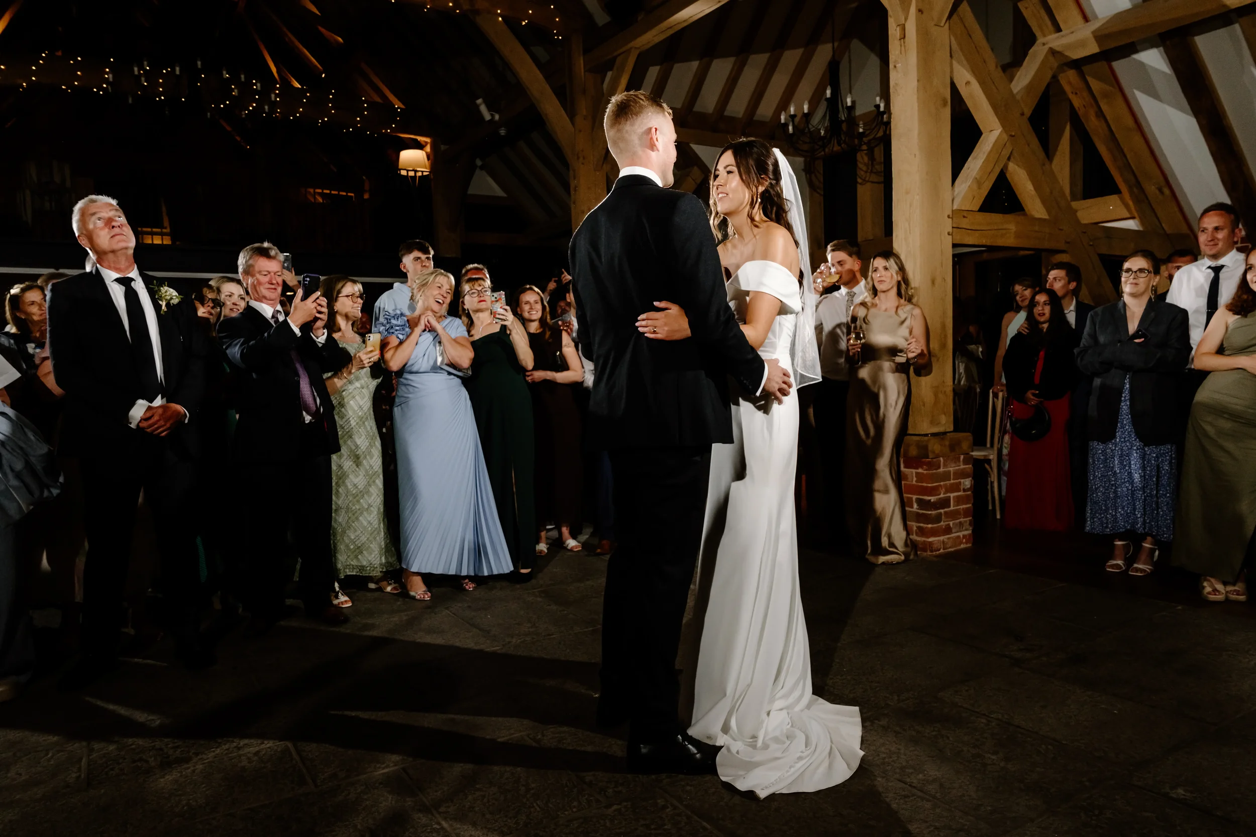 A bride and groom share their first dance at wedding reception, surrounded by guests watching and taking photos inside a rustic venue with wooden beams and warm lighting.