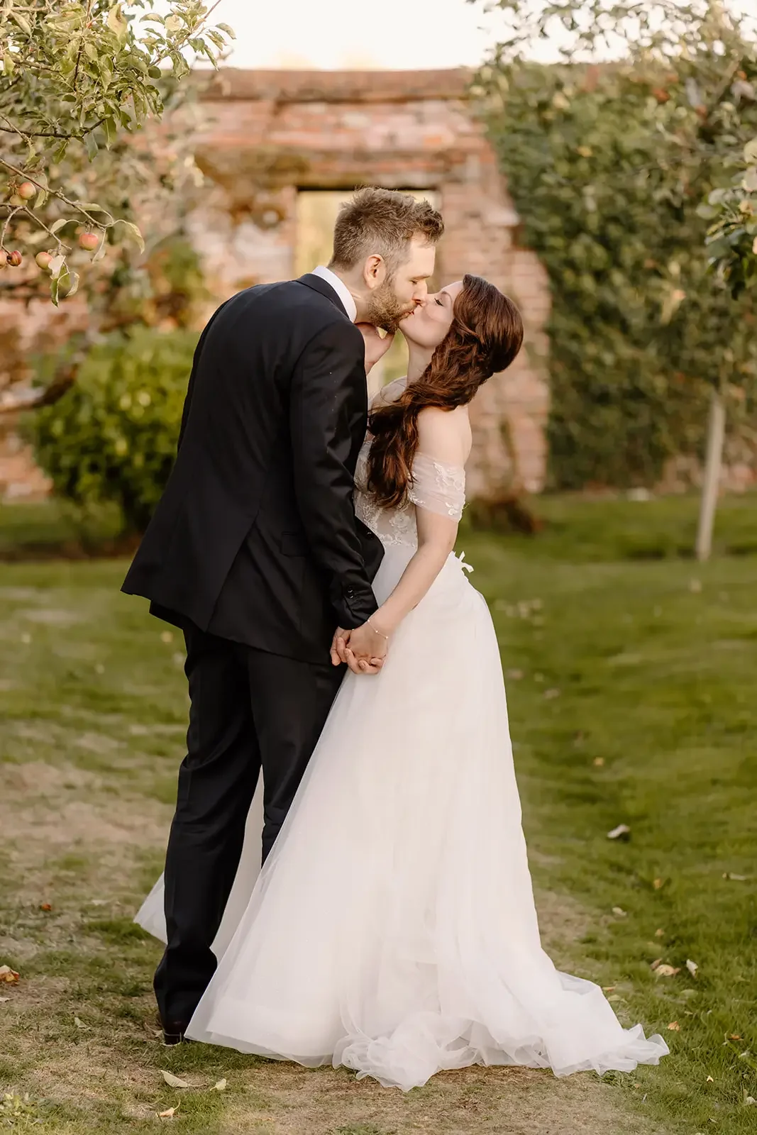 A couple dressed in wedding attire sharing a kiss outdoors in front of an old brick structure and green trees.