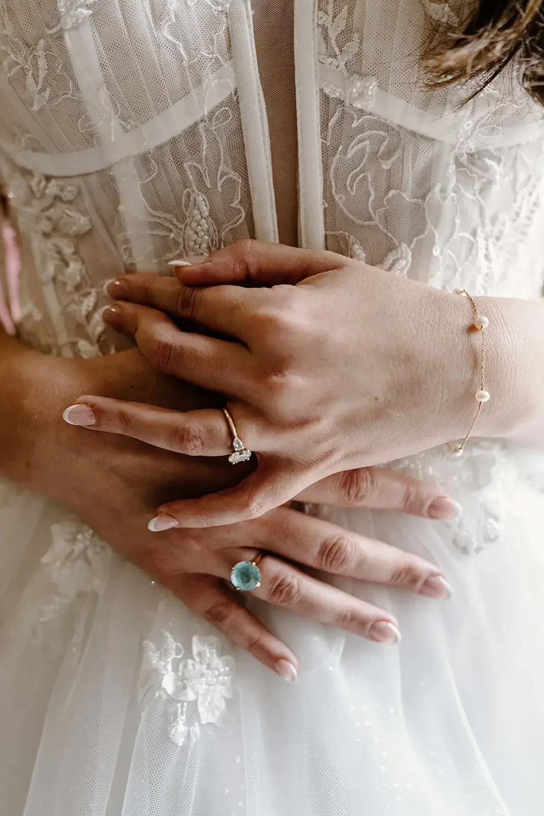 Close-up of two hands with rings, one with a diamond ring and the other with a large aquamarine ring, resting on a bride's lace wedding dress with embroidered floral designs.