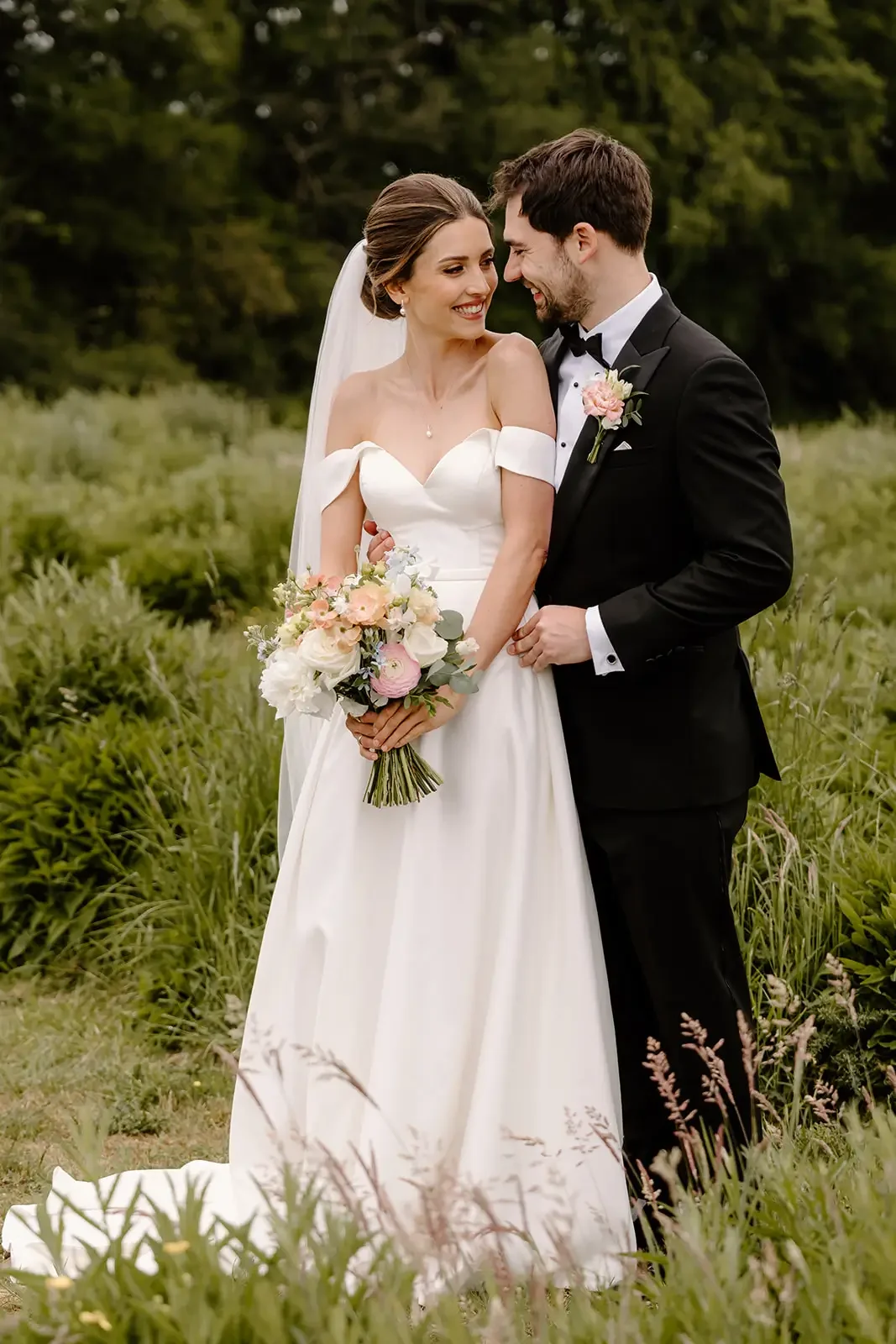 A bride and groom smiling affectionately outdoors in a green field, with the bride holding a bouquet of pink and white flowers. The bride is wearing a white wedding gown with off-shoulder sleeves, and the groom is dressed in a black tuxedo with a pin