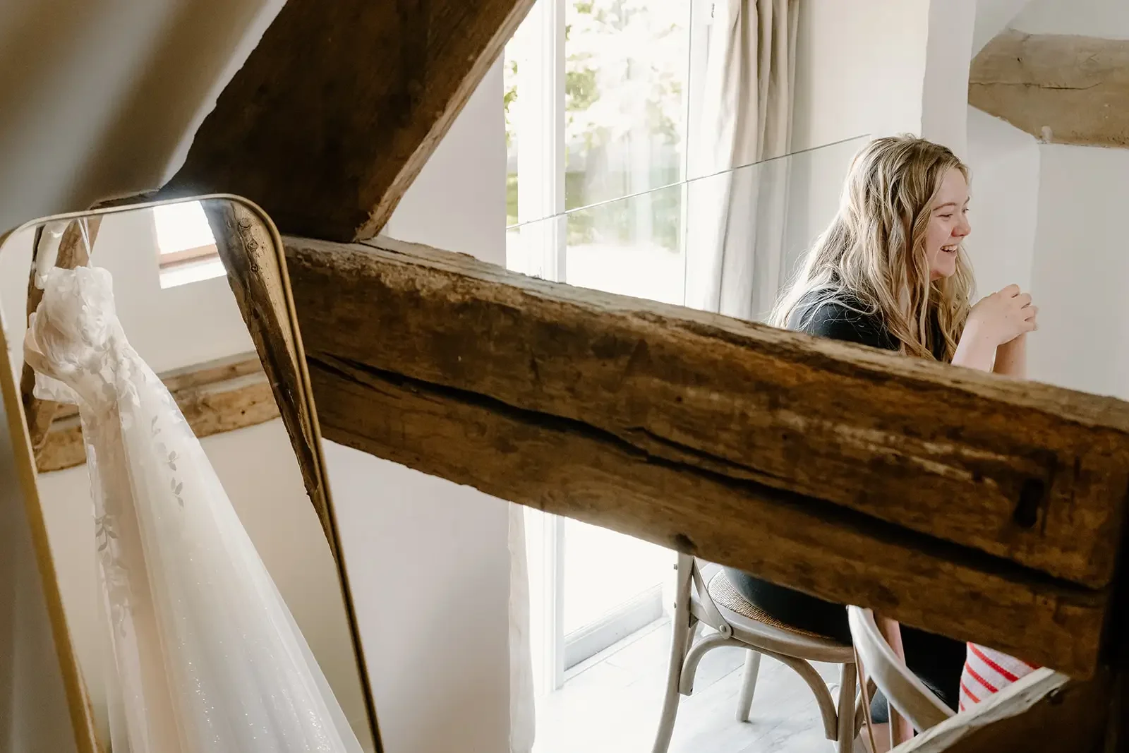 A woman with long blonde hair sitting at a table, smiling, in a room with a large window and wooden beams, with a wedding dress hanging nearby.