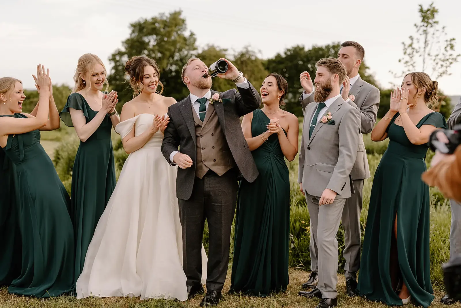 A group of people at a wedding celebration outdoors, with a man in a suit drinking from a bottle surrounded by smiling women and men in formal attire, all clapping and enjoying the moment.