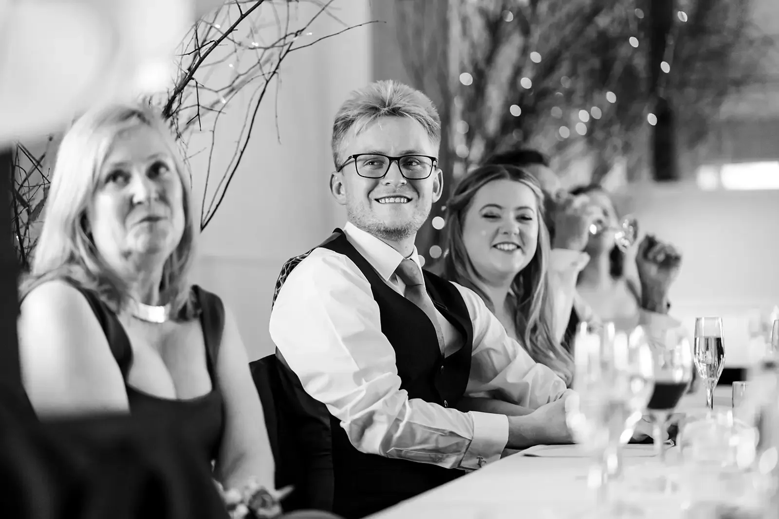 Black and white photo of people sitting at a banquet table, including a man with glasses smiling and two women, one with long hair, all dressed nicely.
