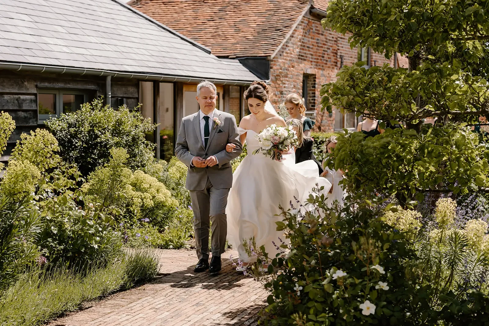 Bride in a white wedding dress holding a bouquet walking arm-in-arm with a man in a gray suit on a garden path with greenery and flowers.