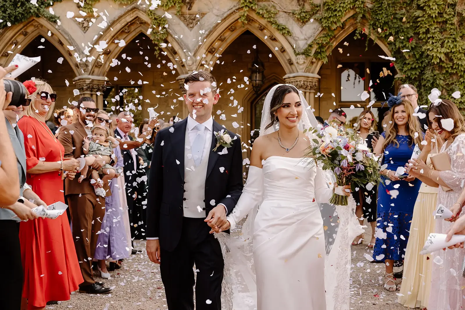 Bride and groom walking hand in hand as guests throw flower petals at their wedding celebration outside a stone building with arches and ivy.