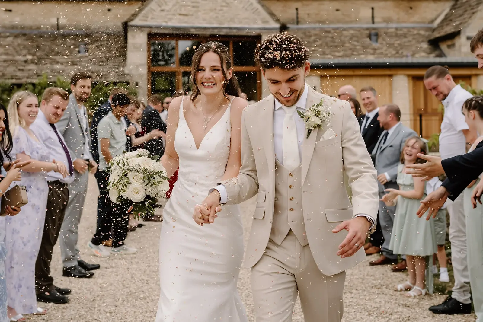 Bride and groom walking hand in hand through a shower of confetti, smiling and joyful, as friends and family celebrate their wedding outside a rustic building.