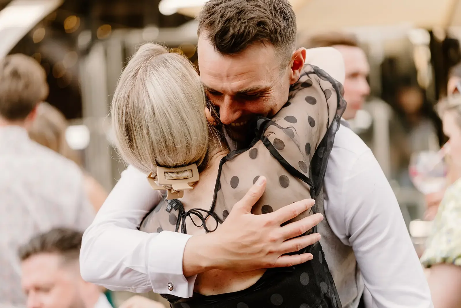 A man and a woman embrace tightly at a social gathering. The man is smiling with his eyes closed, wearing a white shirt, and the woman has blonde hair held back with a clip, wearing a black sheer polka dot dress. Other people are visible in the backg