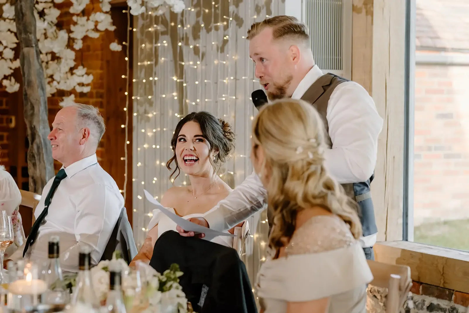 A man giving a speech at a wedding reception, holding a microphone and some papers, with a bride and other guests sitting at a decorated table.