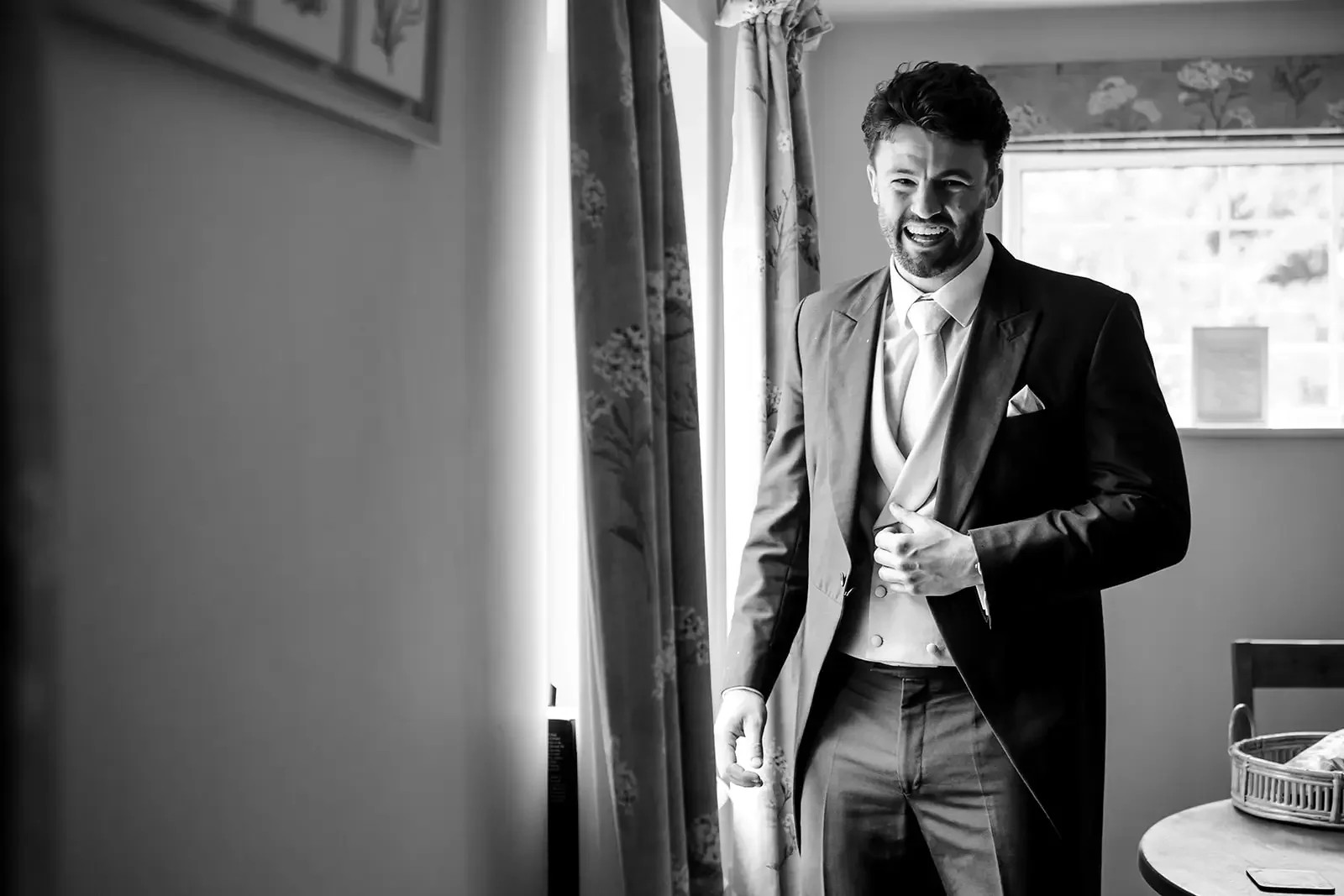 A man in a formal suit smiling and adjusting his jacket in a room with floral curtains and a window.
